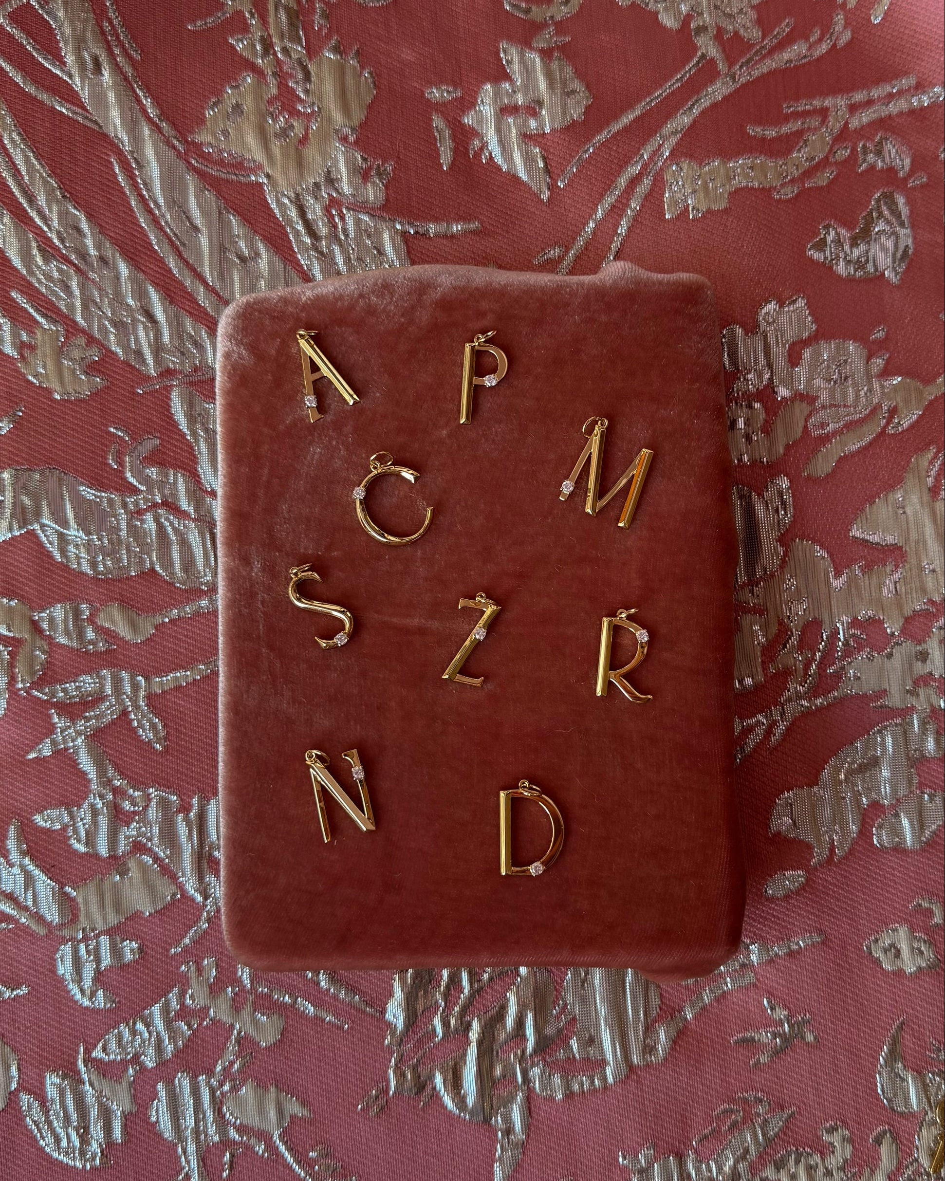 Eloise Letter Charms by Mod + Jo are displayed as gold earrings on pink velvet, with letters A, P, C, M, S, Z, R, and N. Set against a pink and silver textured background—ideal to pair with your favorite gold initial necklace.