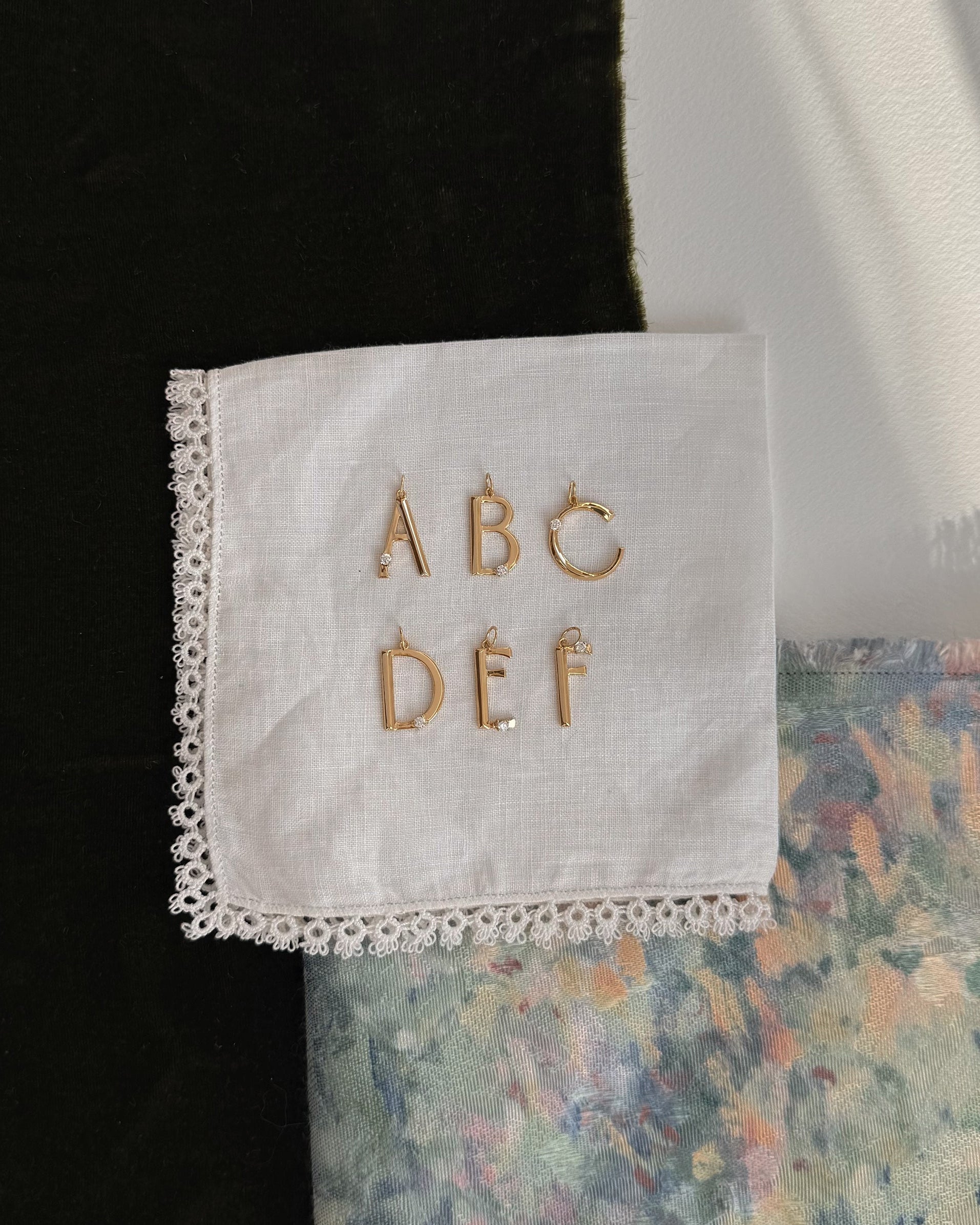 The Mod + Jo Eloise Letter Charm, a gold initial pin, adorns a white lace-edged handkerchief that rests on dark fabric and pastel floral textile as sunlight casts gentle shadows across the scene.
