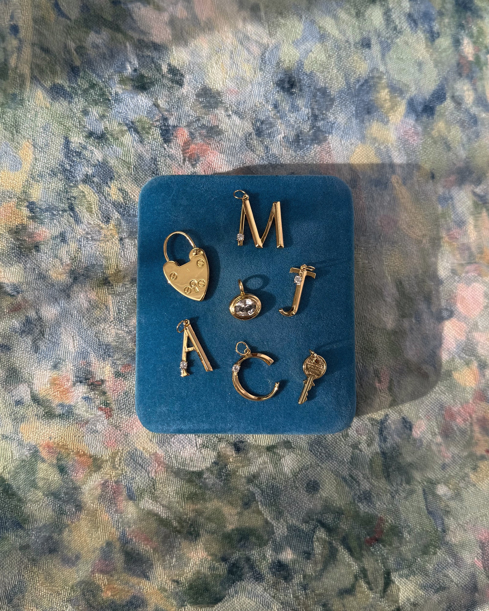 The Eloise Letter Charm by Mod + Jo, available in A, C, J, or M, plus a lock, key, and heart charm—some with sparkling cubic zirconia—are displayed on a blue velvet box over floral fabric.