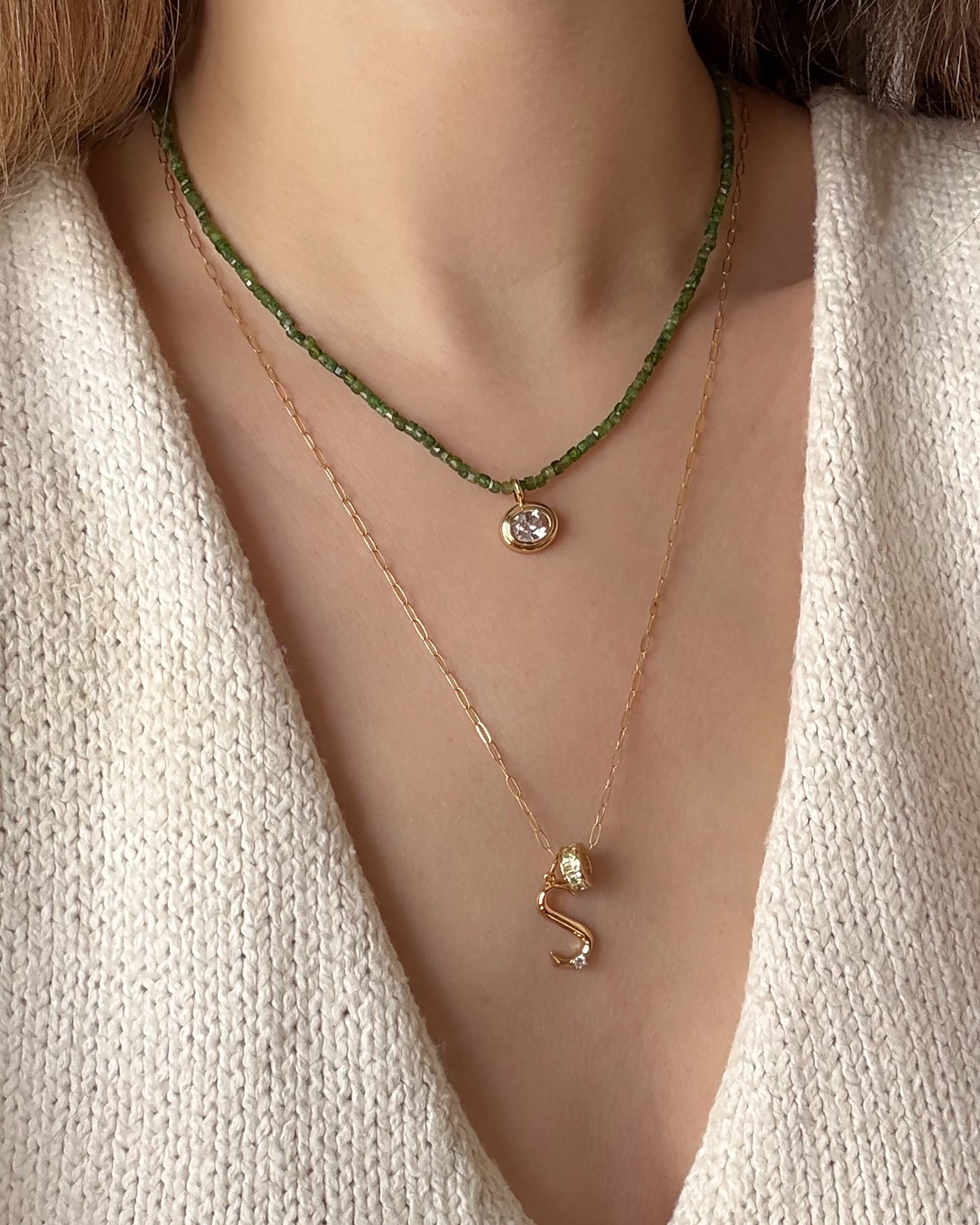 A person in a cream knit V-neck top wears two necklaces: a short green beaded necklace with a round pendant and a longer gold chain with Mod + Jo's Marla Sapphire Bezel Charm—a 14k gold S-shaped pendant with lab-grown sapphire.