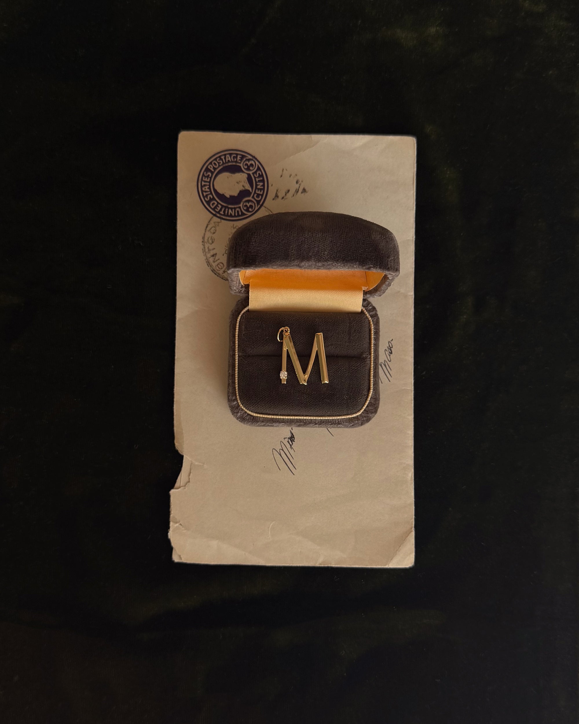 The Eloise Letter Charm by Mod + Jo, a gold "M" initial brooch with a small pearl accent, is displayed in a brown velvet box with yellow lining atop a vintage envelope featuring handwritten signatures and a postal stamp.