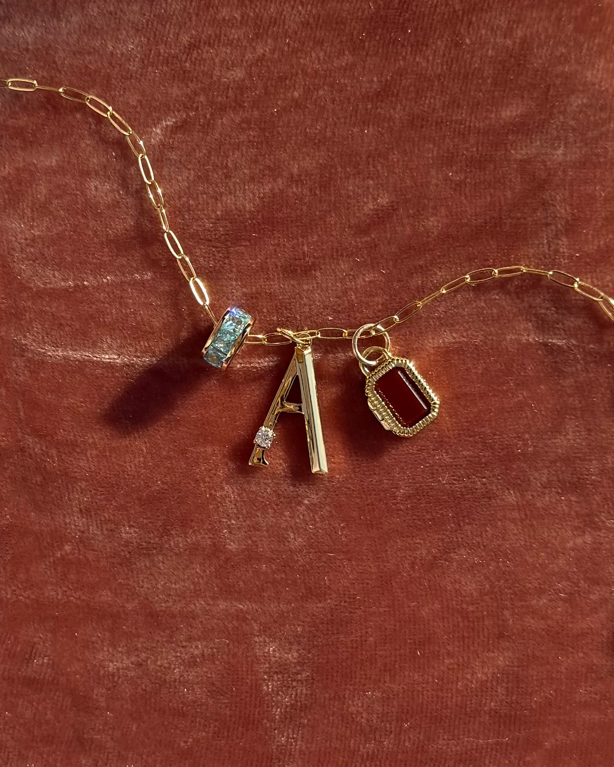 The Eloise Letter Charm by Mod + Jo is a gold plated chain with three charms—a clear gemstone, initial A charm, and red gemstone in a gold frame—displayed on reddish-brown velvet fabric.