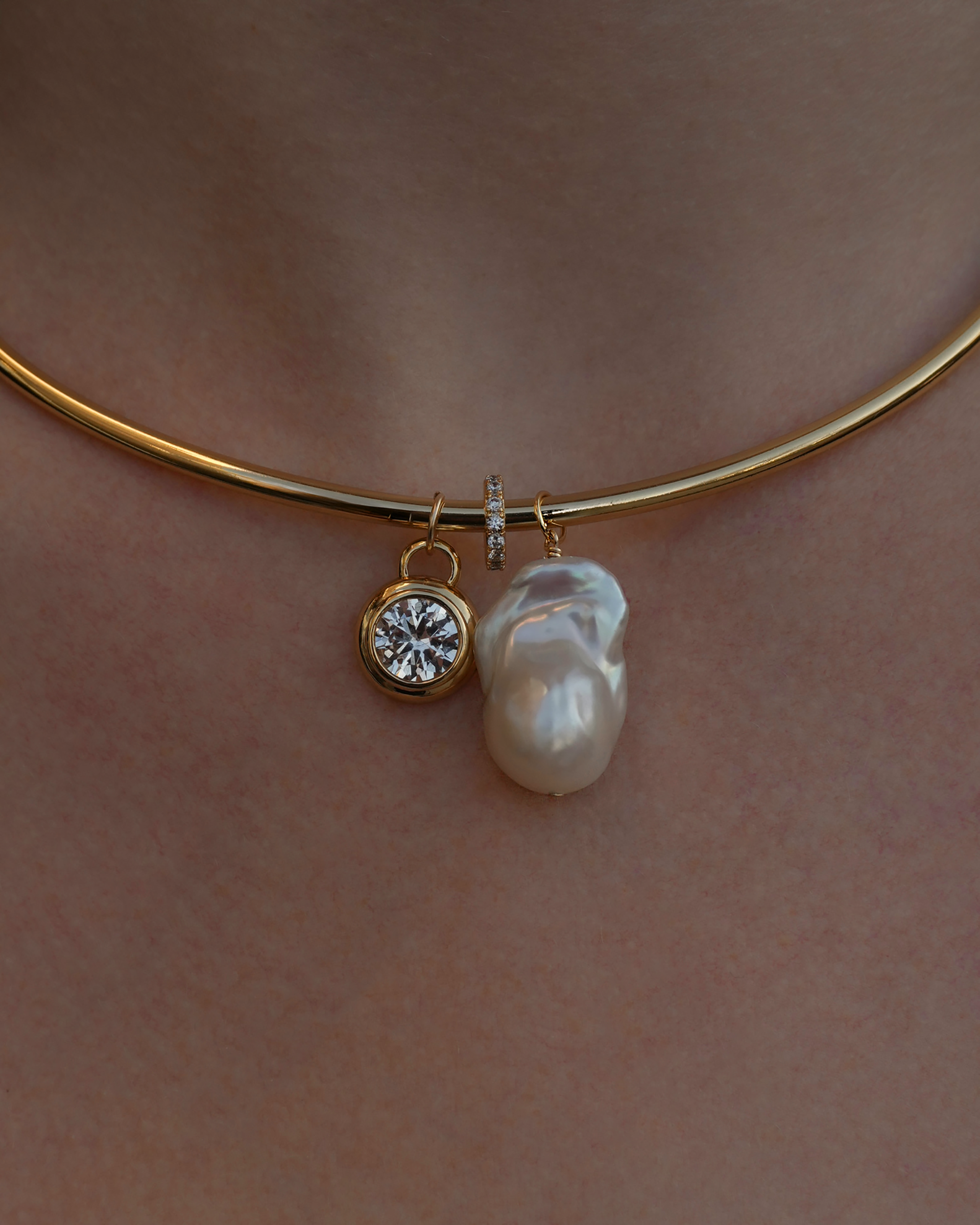A close-up of a gold choker necklace featuring three pendants: a round diamond, a small gold ring with tiny diamonds, and a large, irregularly shaped pearl, worn on a persons neck.