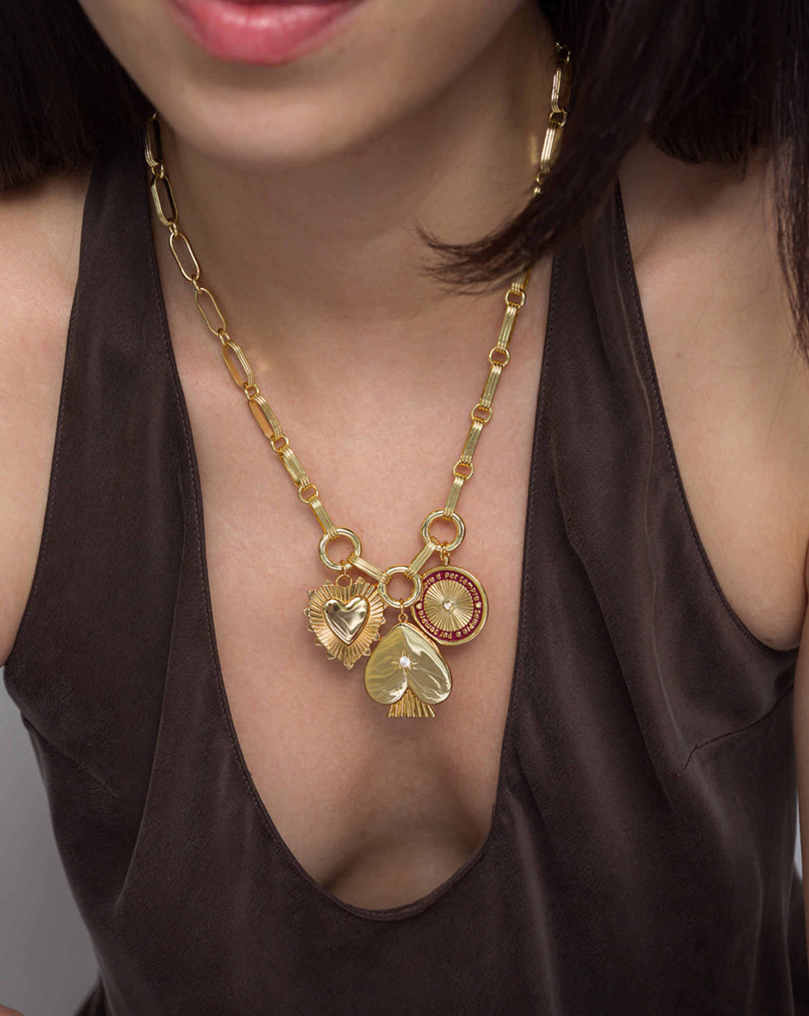 A person wearing a brown V-neck top and the Mod + Jo Jagger Charm Clip Necklace, a chunky 14k gold piece featuring three pendants: a heart, a leaf, and a round charm with red accents.
