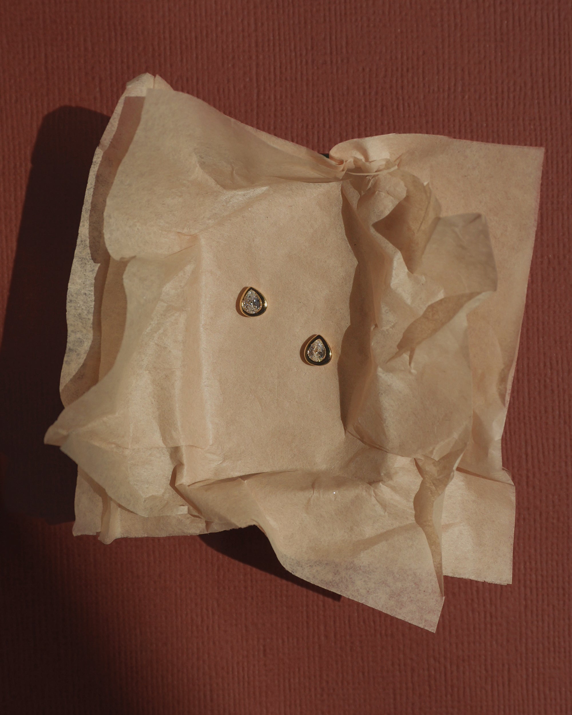 Joni Bezel Studs by Mod + Jo, featuring two small gold-plated teardrop-shaped earrings with a clear cubic zirconia center, rest on crumpled beige tissue paper against a textured brown background, highlighting their subtle elegance.