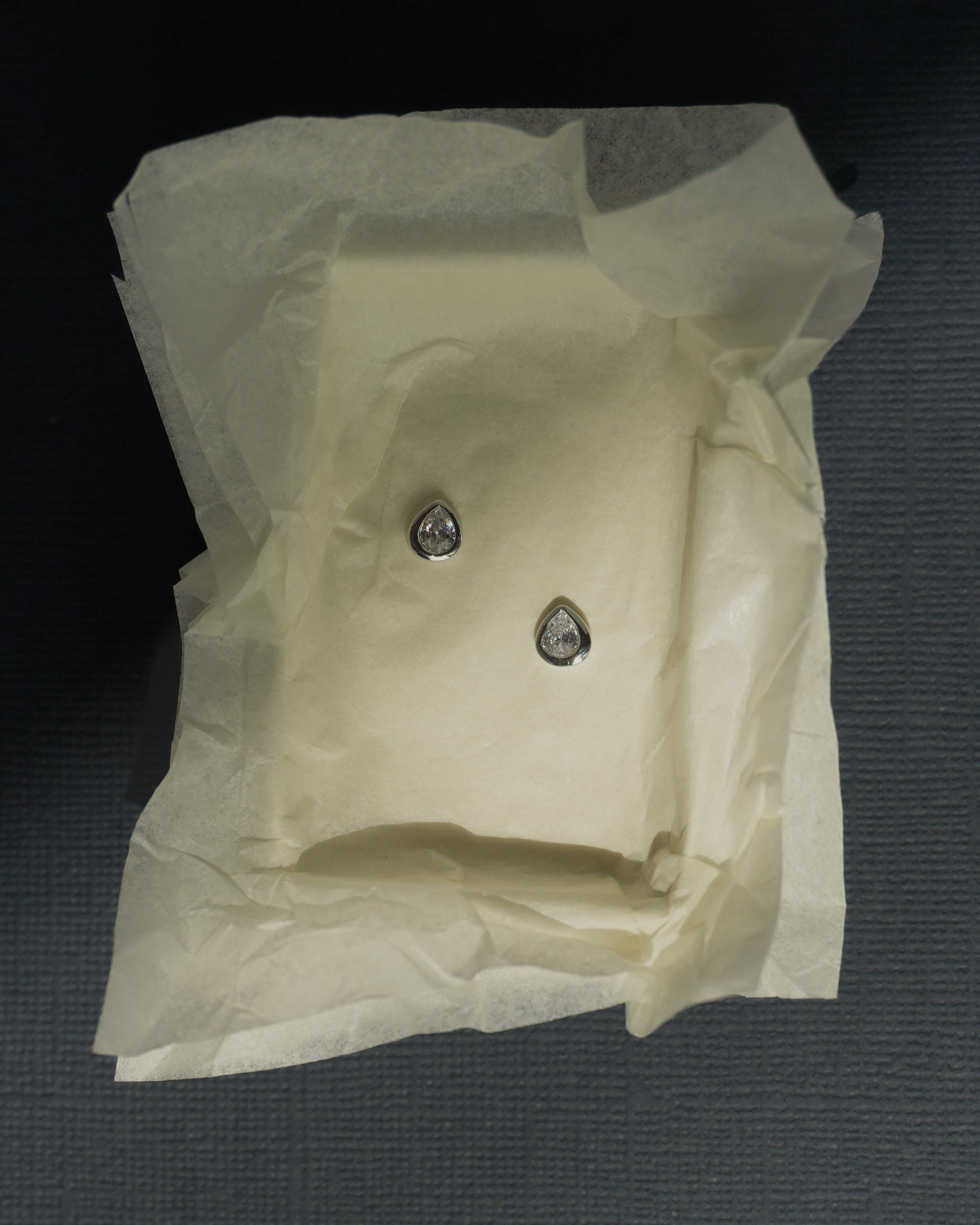 A pair of small, pear-shaped diamond earrings rests on crumpled white tissue paper inside a box, their brilliance reminiscent of the elegant Joni Bezel Studs by Mod + Jo.