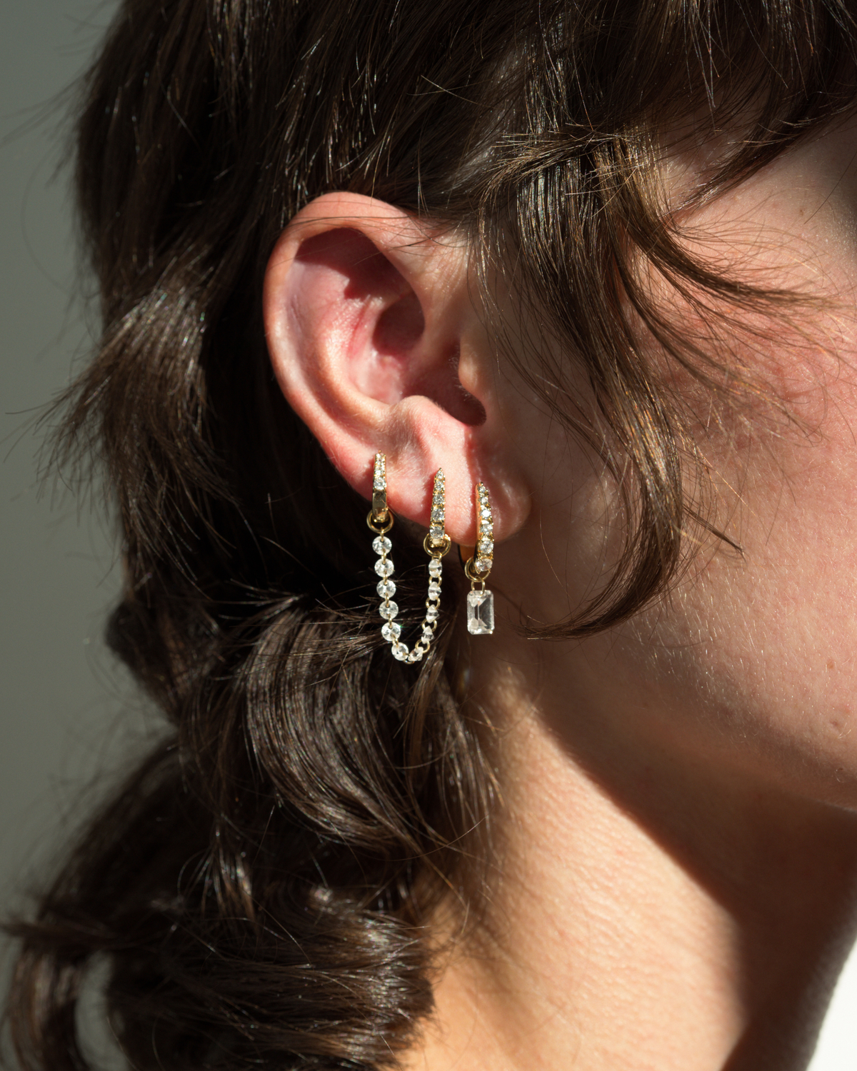 Jules Grande Huggies featuring two 14k gold plated hoop earrings connected by a delicate chain of cubic zirconia crystals, with a third hoop showcasing a dangling emerald-cut crystal.