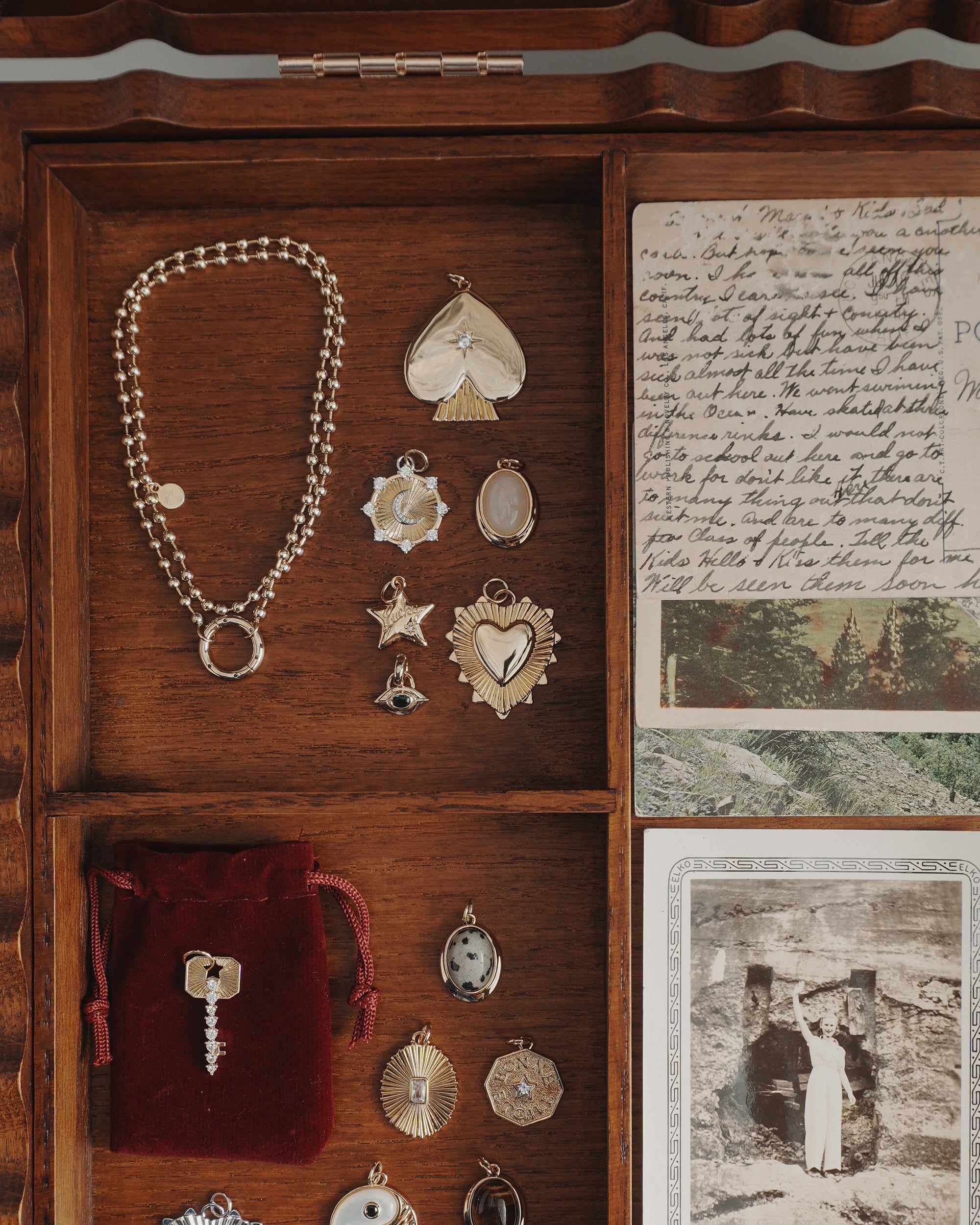 A wooden jewelry box contains gold charms, a bracelet, a protective Mod + Jo Nyx Evil Eye Charm pendant, a red velvet pouch holding a key, vintage postcards, and a black-and-white photograph of a person standing outside against a stone wall.
