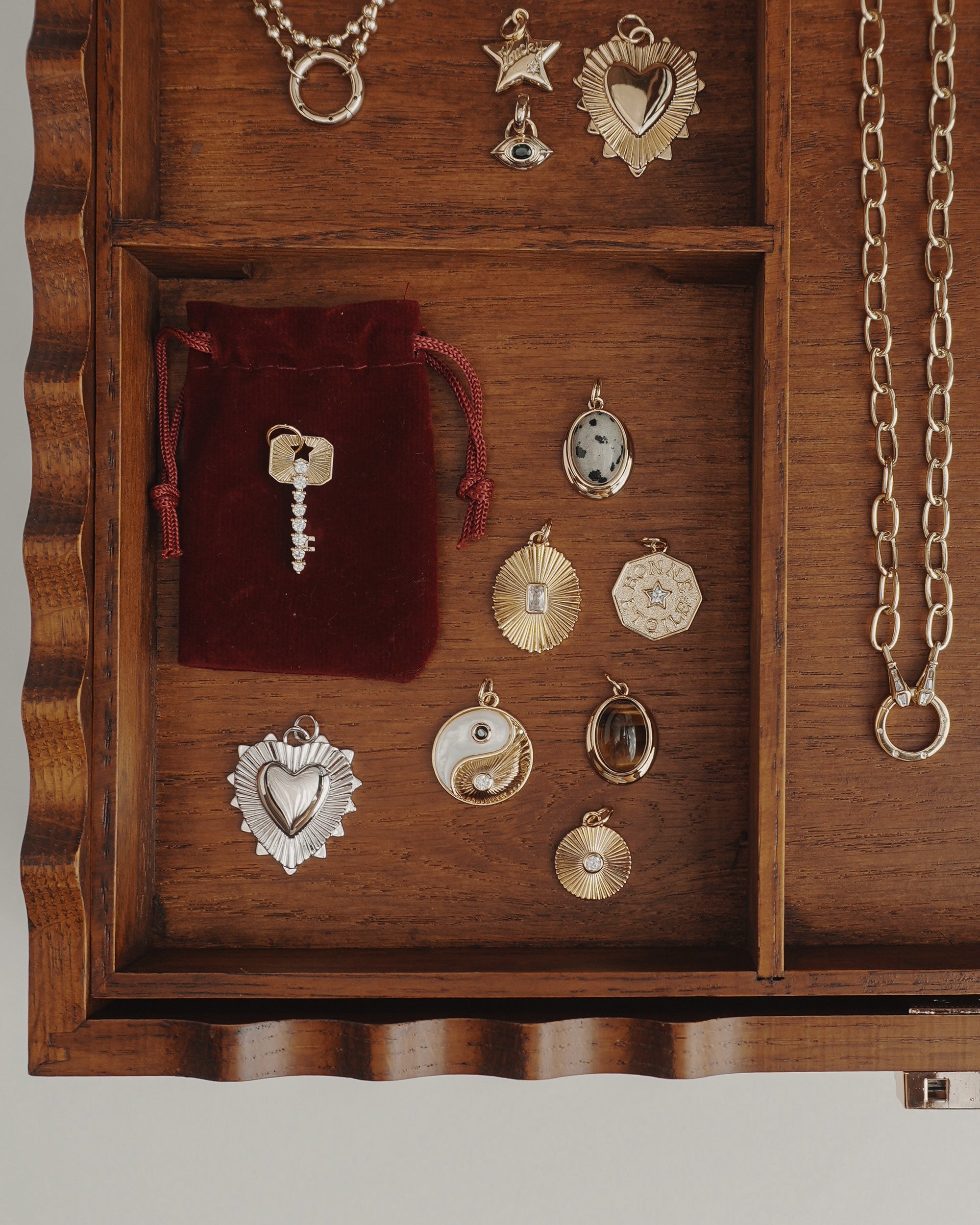 A wooden jewelry box featuring elegant 14k gold jewelry and an assortment of gold and silver pendants, chains, and a red velvet pouch. In the neatly organized compartments are various charms—including hearts, stars, an eye, and the Mod + Jo Grande Lynn Engravable Heart Charm—crafted for personalization.