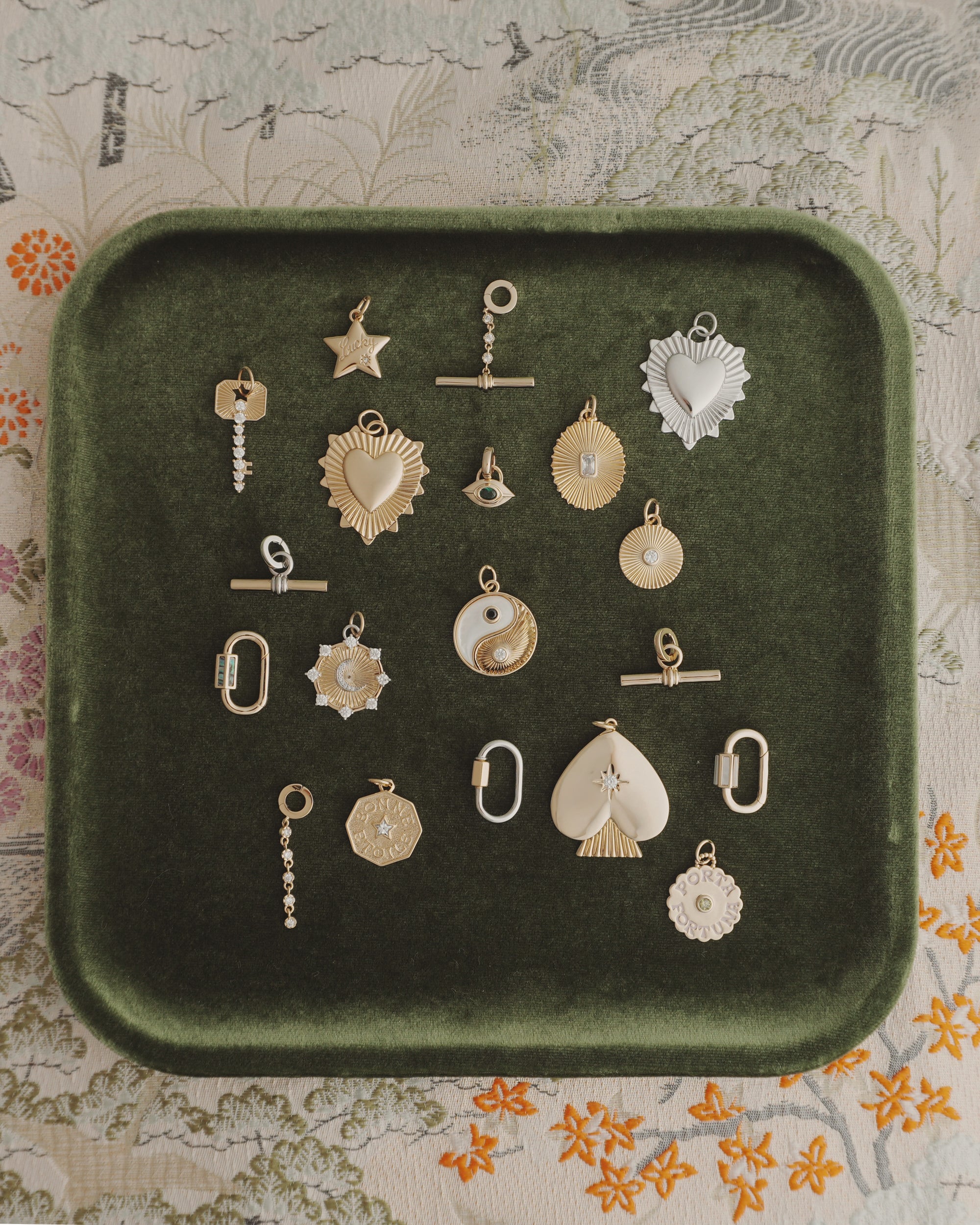 A Nora Lock Carabiner by Mod + Jo is displayed on a green velvet tray alongside 18 assorted gold and silver charms with 14k plating—featuring hearts, stars, circles, a fan, yin-yang, and geometric designs—all arranged on a floral-patterned fabric background.