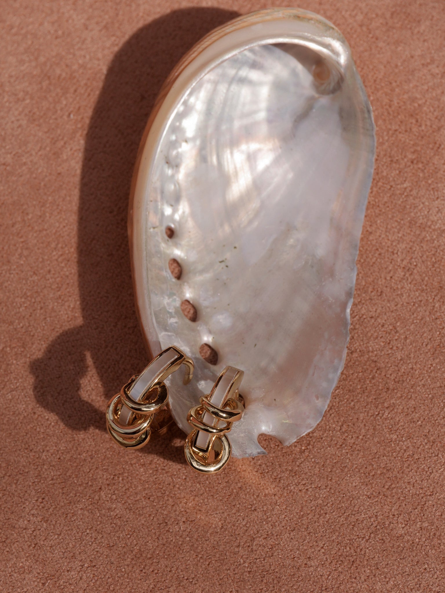 The Mod + Jo Levi Huggies, featuring interlocking gold loops, are displayed in front of a large iridescent seashell on a textured light brown surface.