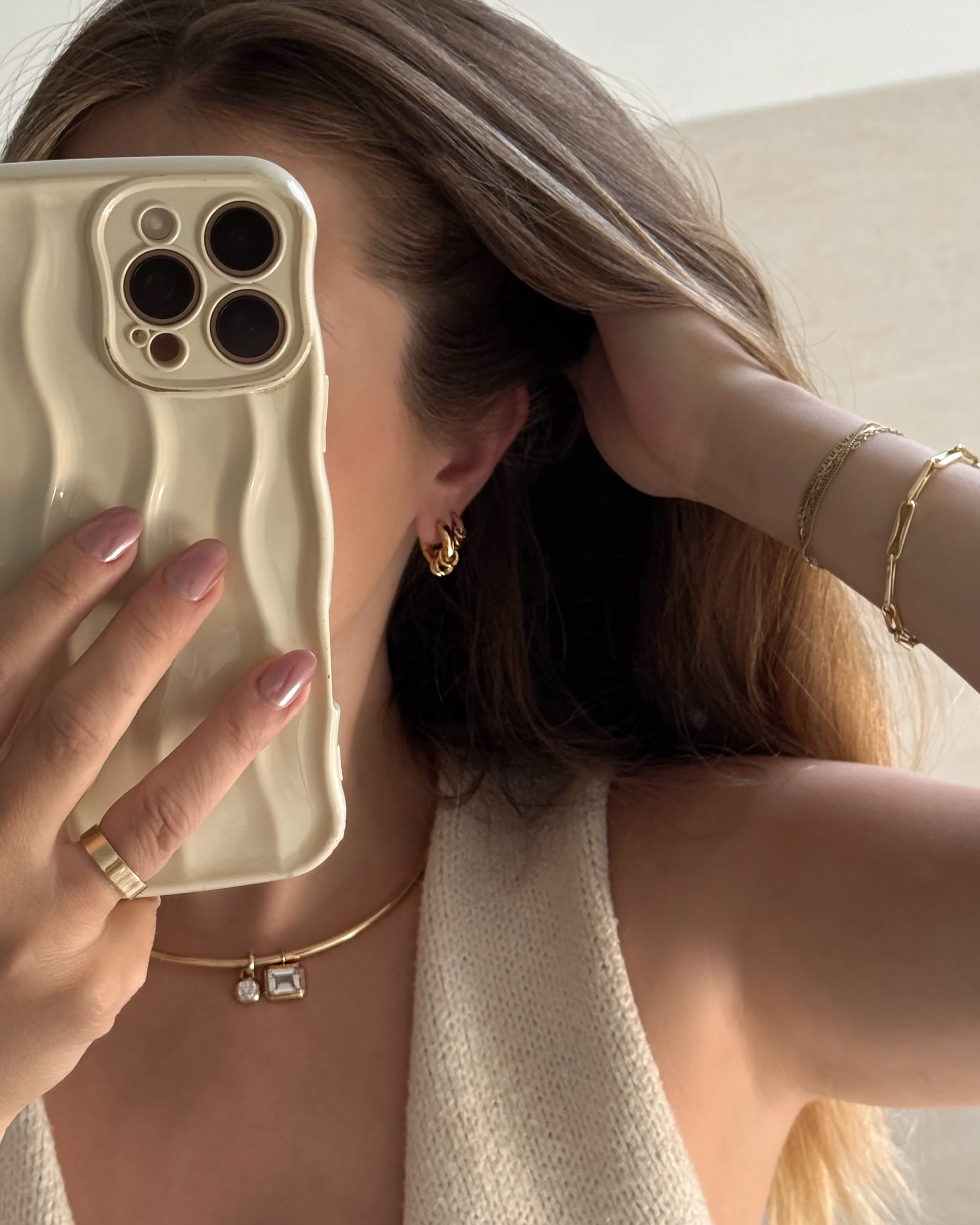 A woman with long hair takes a mirror selfie, her face hidden behind a cream-colored wavy phone case. She accessorizes with gold jewelry—hoop earrings, a necklace, and a ring—paired with a sleeveless knit top. On her wrist, she stacks the Mod + Jo Molly Elongated Link Chain Bracelet alongside other layered bracelets for an effortlessly chic look.