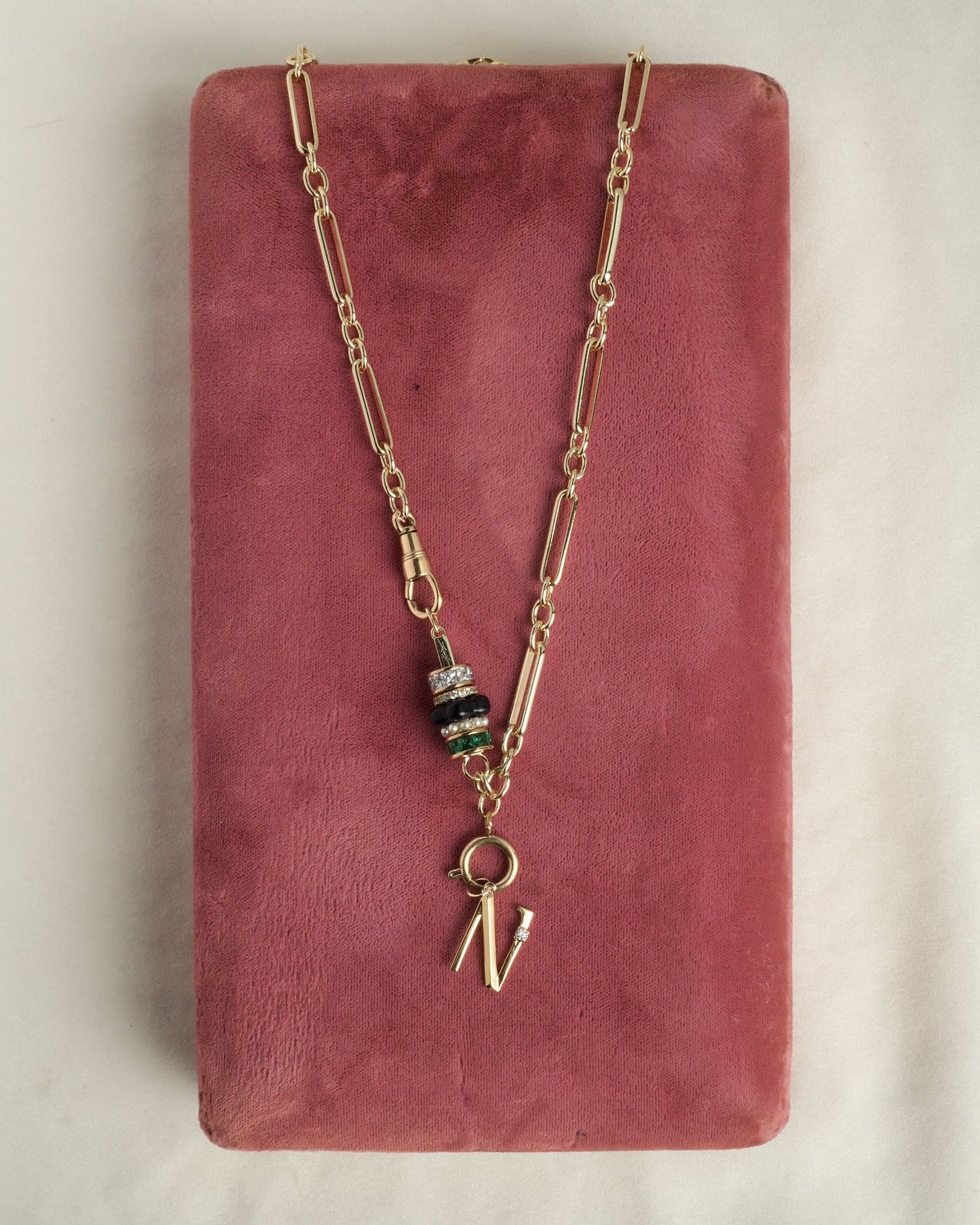 The Eloise Letter Charm by Mod + Jo features a gold initial necklace with paperclip links, stone beads, and a sparkling cubic zirconia "N" charm, elegantly displayed on a rose-colored rectangular fabric surface.