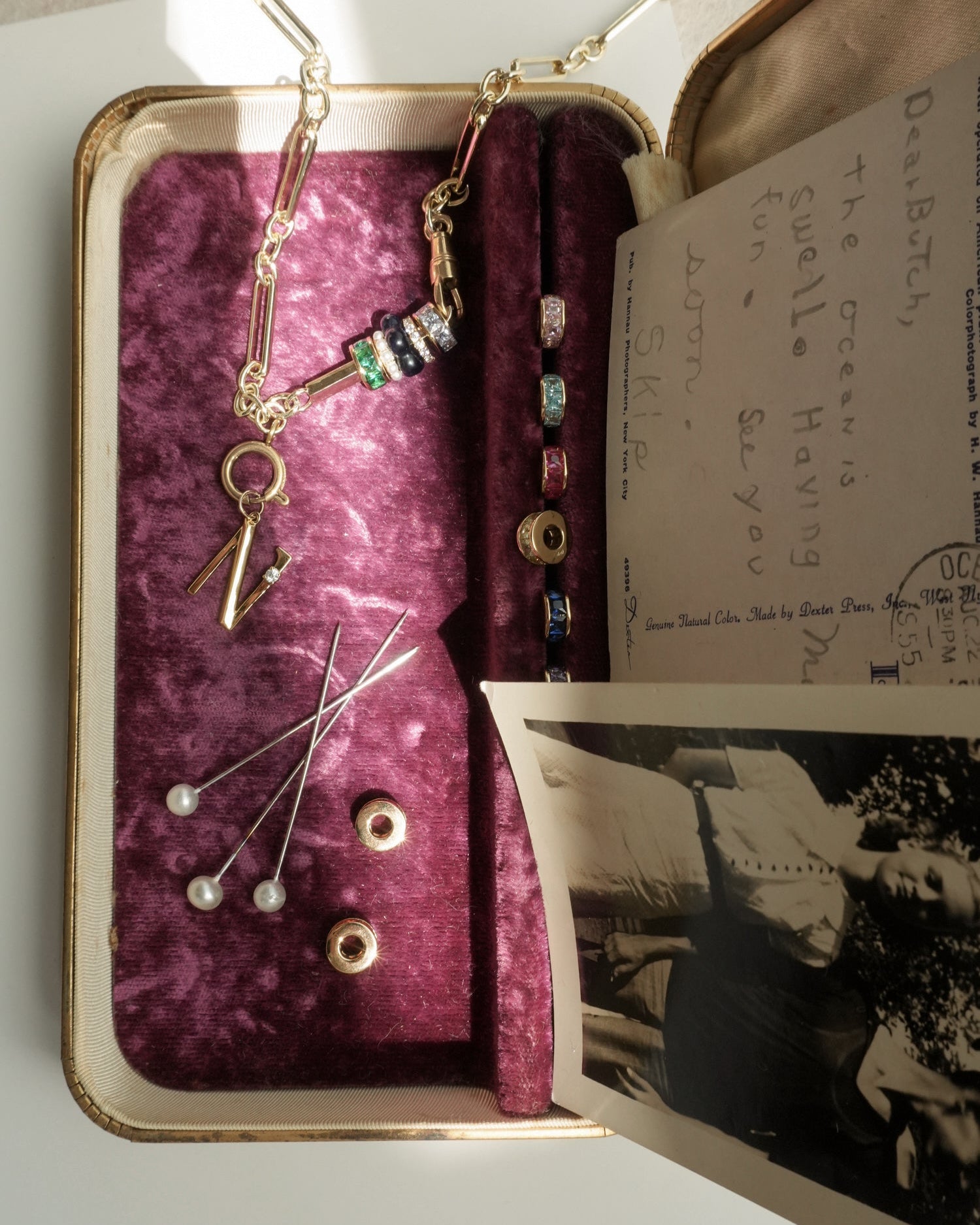 An open velvet-lined jewelry box features the Mod + Jo Eloise Letter Charm necklace, colorful beads, pearl hatpins, loose beads, and letter charms. A black-and-white photo and vintage postcard inside are lit by sunlight.