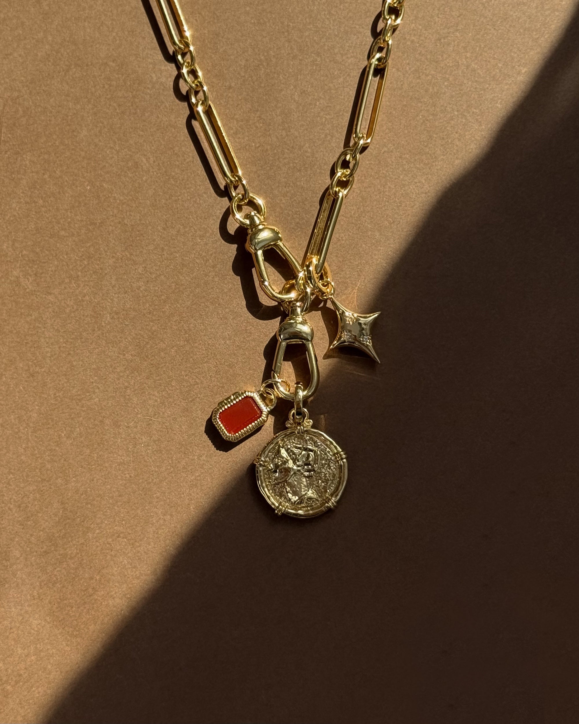 The Mod + Jo Mabel Rectangle Birthstone Locket Charm is a 14k gold plated paperclip chain necklace with four interchangeable charms—a red rectangular gem, coin, crescent moon, and star—shown on a brown background with partial shadow.