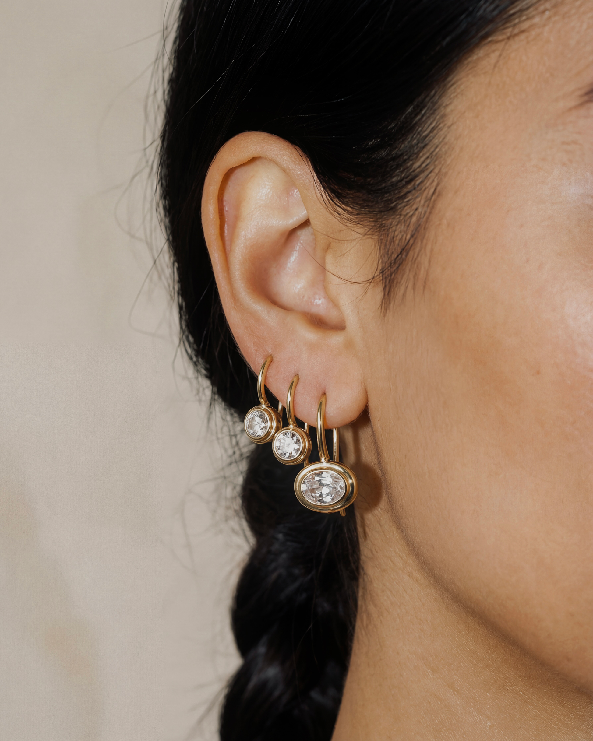 Close-up of an ear wearing gold hoop earrings with clear gemstones on a neutral background
