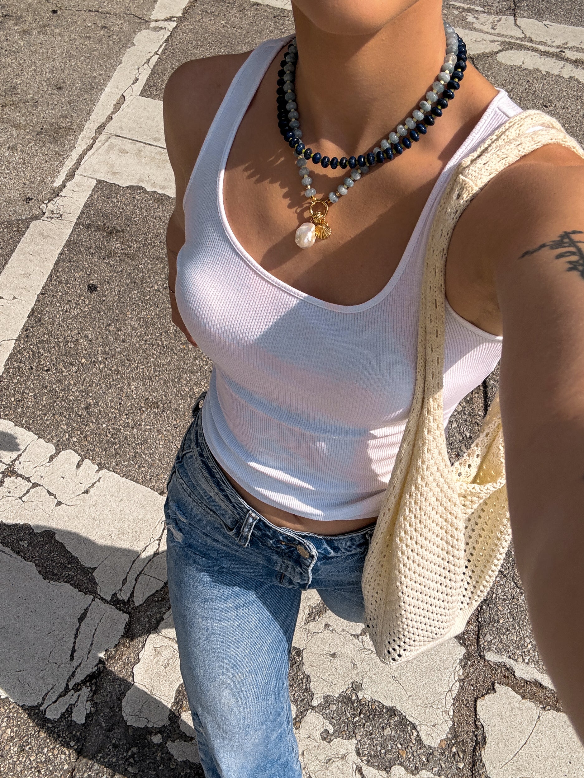 Wearing a white ribbed tank top, blue jeans, and a crochet shoulder bag, a person stands on a sunlit crosswalk. They accessorize with the Mod + Jo Macey Gemstone Necklace and display a partial arm tattoo.