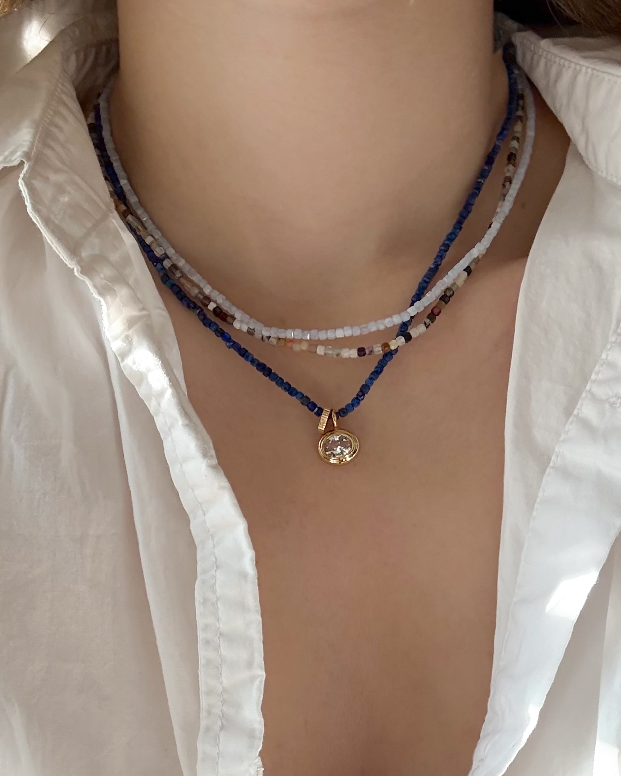 A close-up of a person wearing a white shirt and three layered necklaces: two beaded strands in blue and white, and Mod + Jo's handmade Merit Gemstone Necklace in 14k gold-fill with a clear stone pendant, crafted in Dallas.