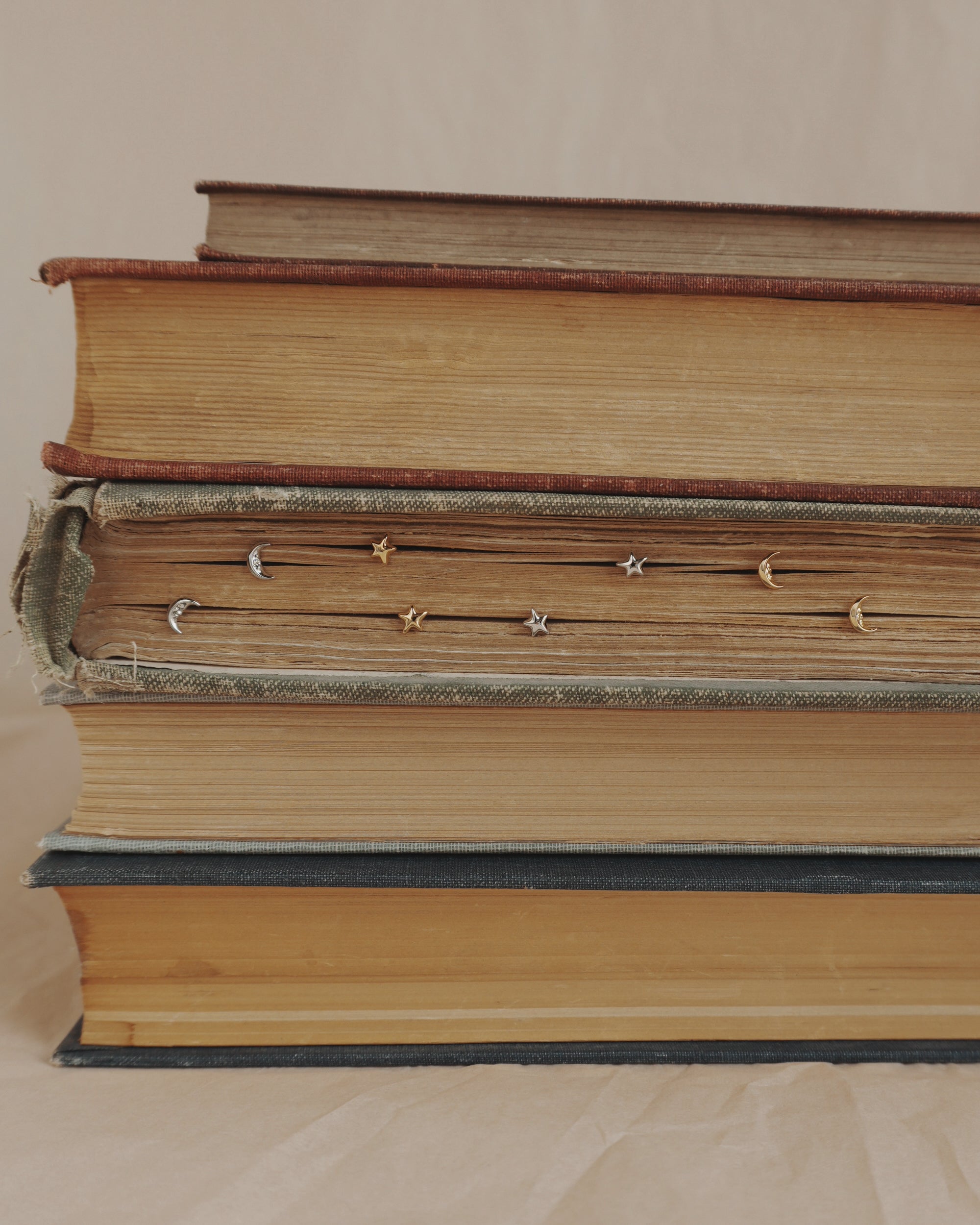 A stack of four old books lies horizontally, with Mod + Jo’s Archie Moon Studs inserted along the edges of the pages in the second book from the top. The background is neutral and plain.