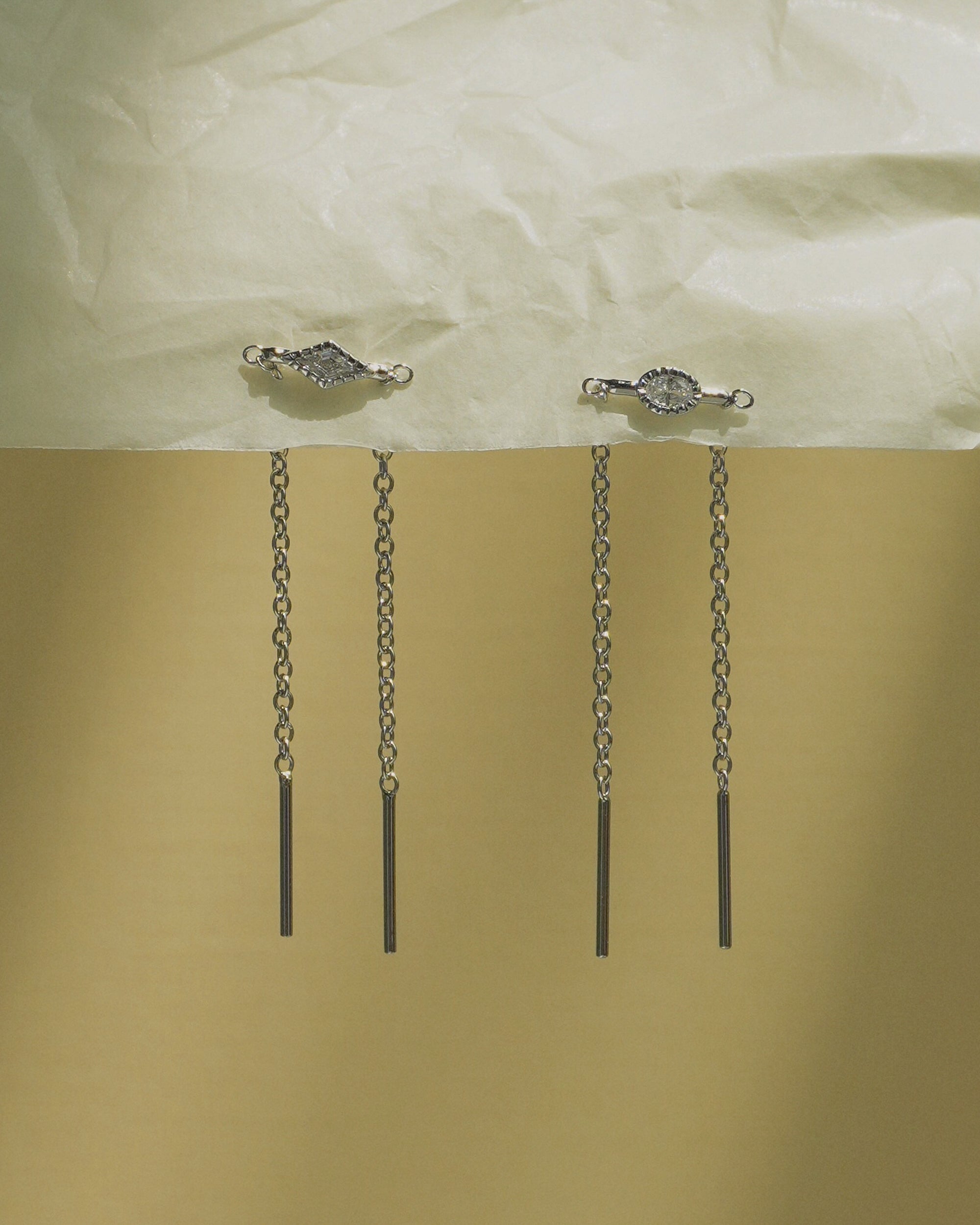 Two Nicole Kite Threader Earrings by Mod + Jo, with geometric and round tops, hang from a textured pale surface. Each earring features two delicate dangling chains ending in straight bars, set against a gold and cream background.