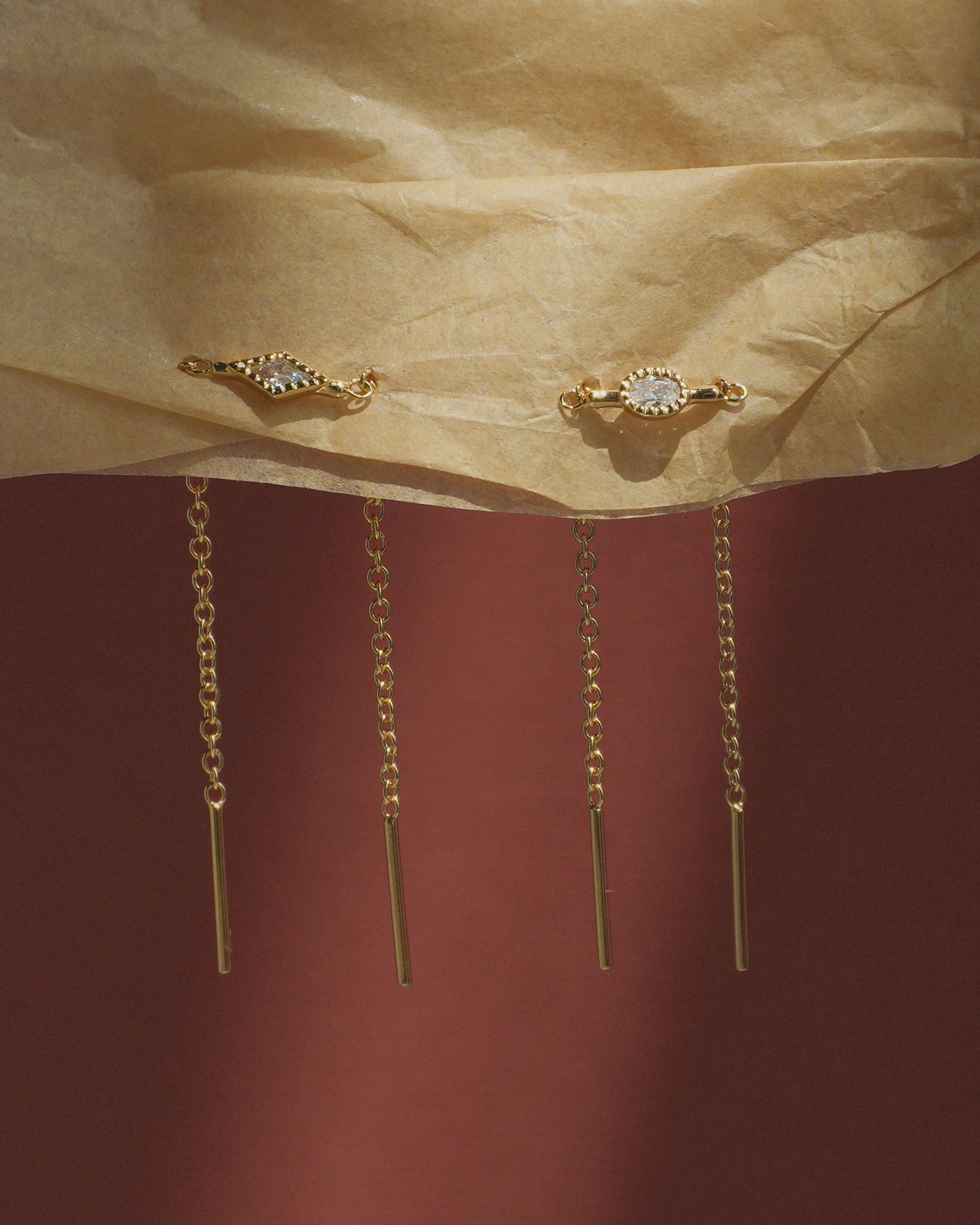 The Nicole Kite Threader Earrings by Mod + Jo, featuring delicate gold-plated chains and a sparkling cubic zirconia stone set in a gold frame at the top, hang against crumpled beige paper with a dark red background.