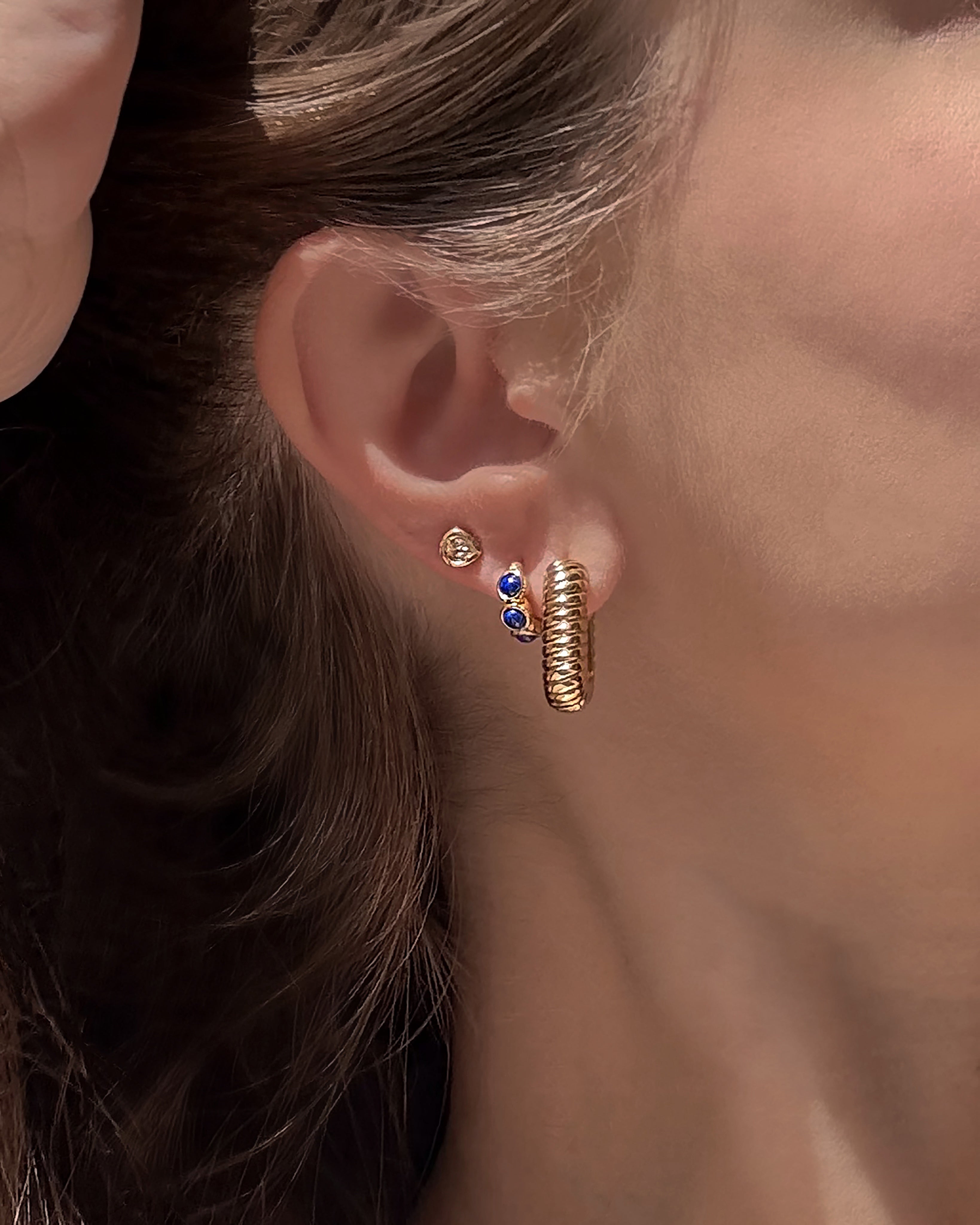 A close-up of a woman’s ear showcases three gold plated earrings from Mod + Jo: a small stud, the Joni Bezel Studs featuring double blue gems, and a textured gold hoop. Her brown hair is neatly tucked behind her ear.