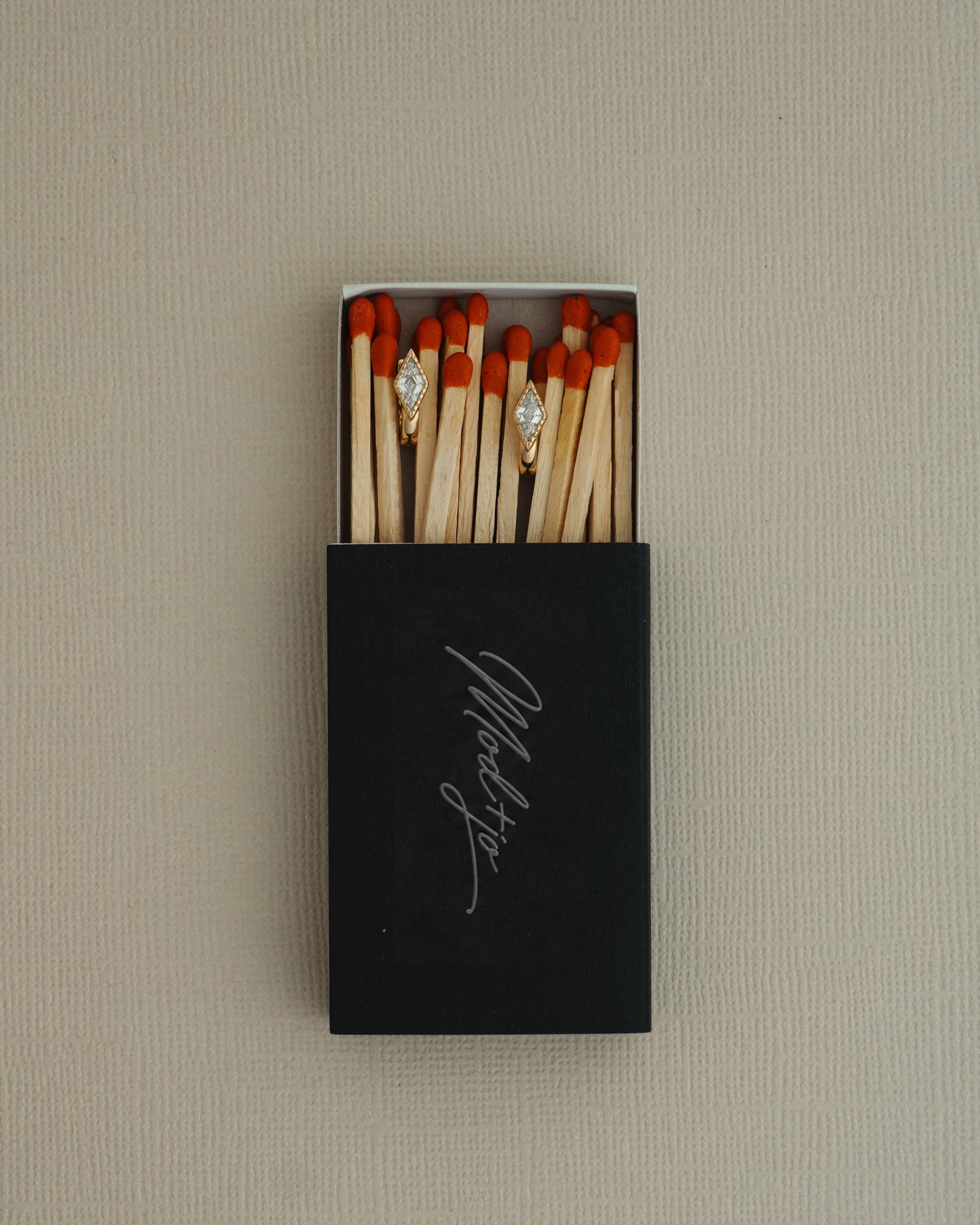 A black matchbox with “Marlsto” written on it, filled with matches, displays two matches adorned with sparkling Phoebe Kite Huggies by Mod + Jo—each shining like dazzling cubic zirconia earrings—set against a neutral textured background.