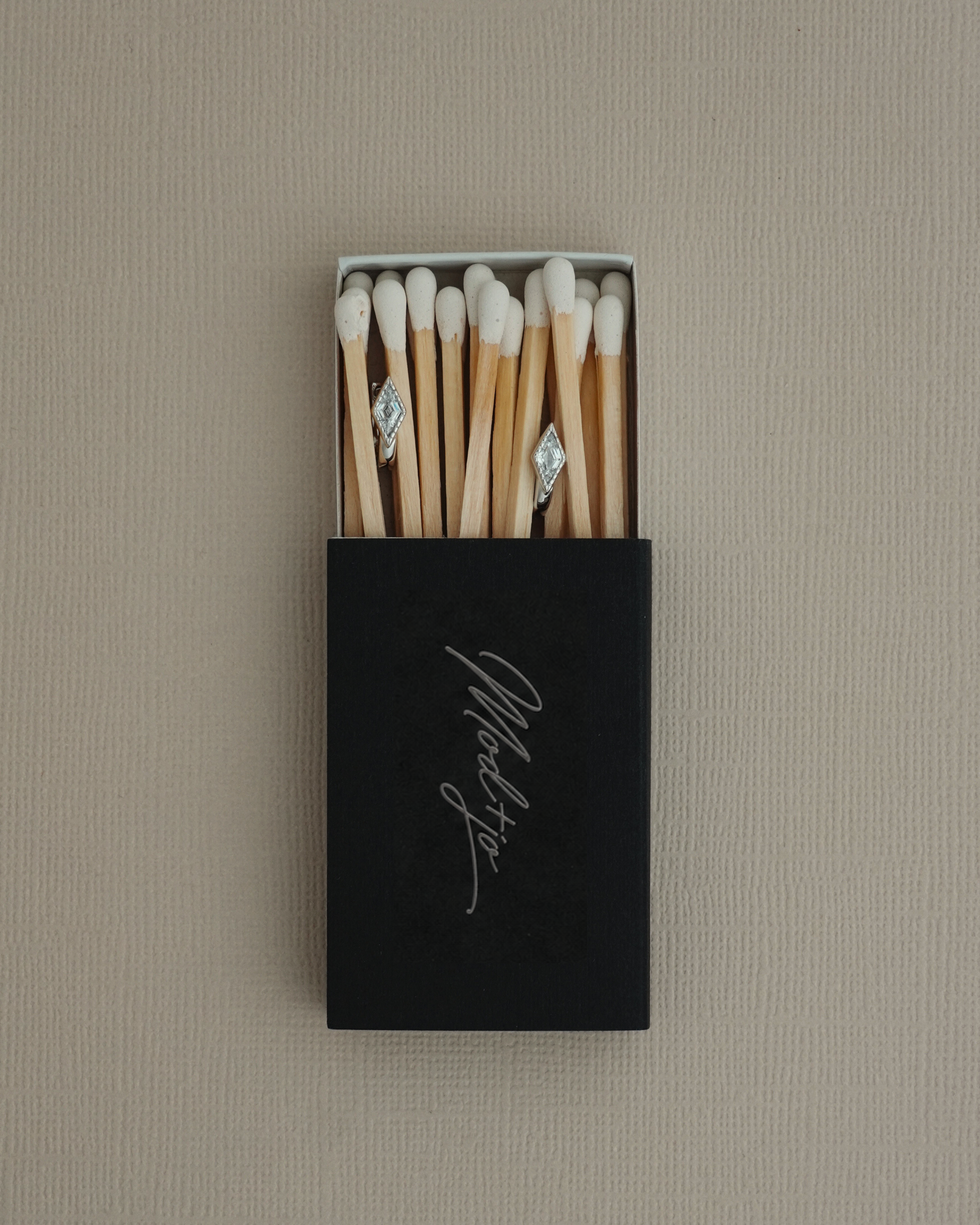 A black matchbox lies open on a textured beige surface, revealing white-tipped matches inside—two of which display sparkling Mod + Jo Phoebe Kite Huggies perched atop their heads like brilliant cubic zirconia.