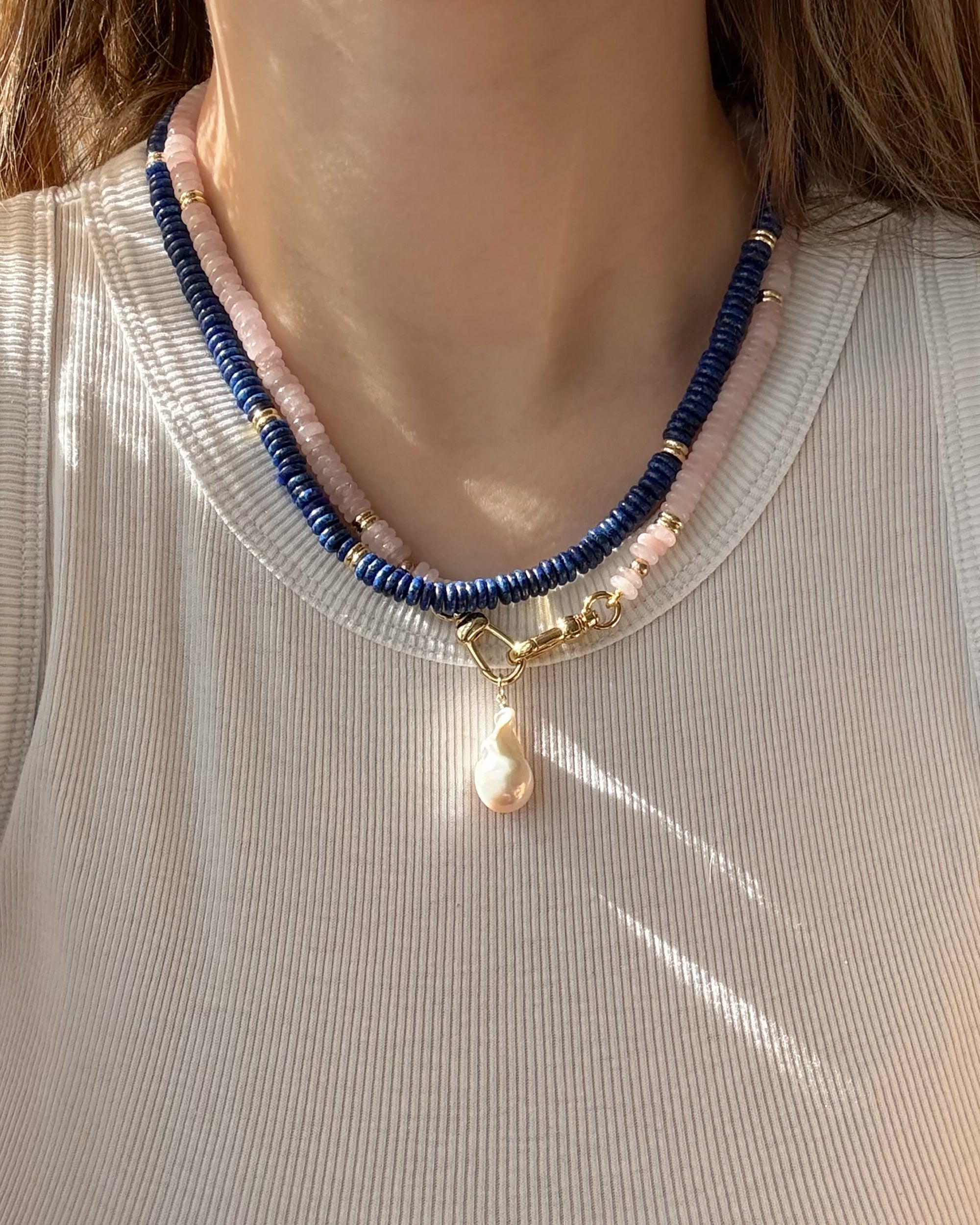 A person wearing a ribbed, light top and the Mod + Jo Sonia Gemstone Necklace—featuring two strands of lapis lazuli and pale pink beads with gold accents and a central pearl pendant.