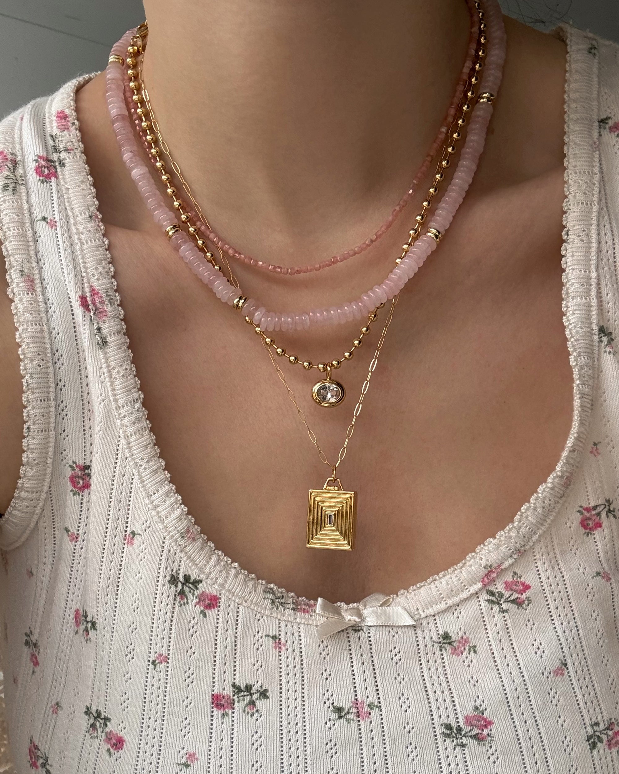 A person wears a white, floral-patterned tank top with a small bow at the neckline. They layer pink bead and gold chain necklaces, including the Mod + Jo Sonia Gemstone Necklace featuring genuine gemstone beads and a rectangular gold pendant.