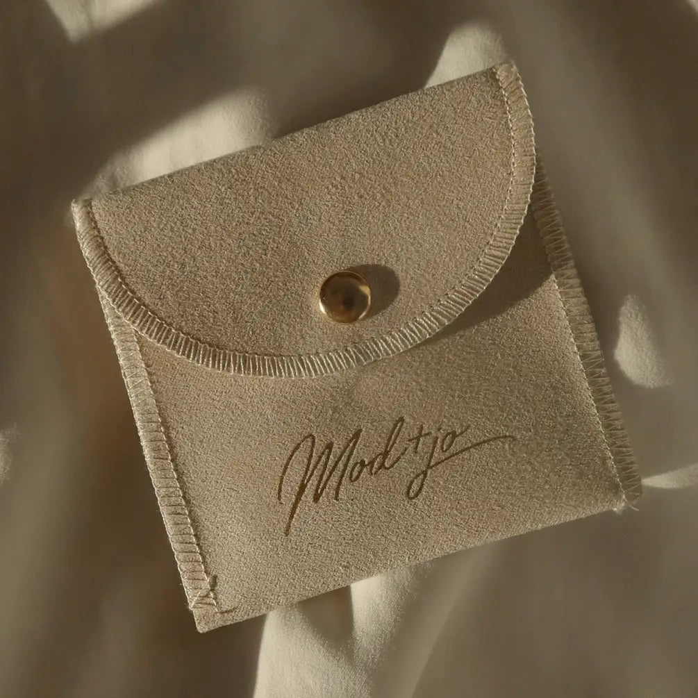 This elegant Mod + Jo Pouch features beige suede, a button closure, and “Modtijo” scripted on the front, making it perfect for storing your favorite jewelry. The pouch rests gracefully on a soft, cream-colored fabric background.