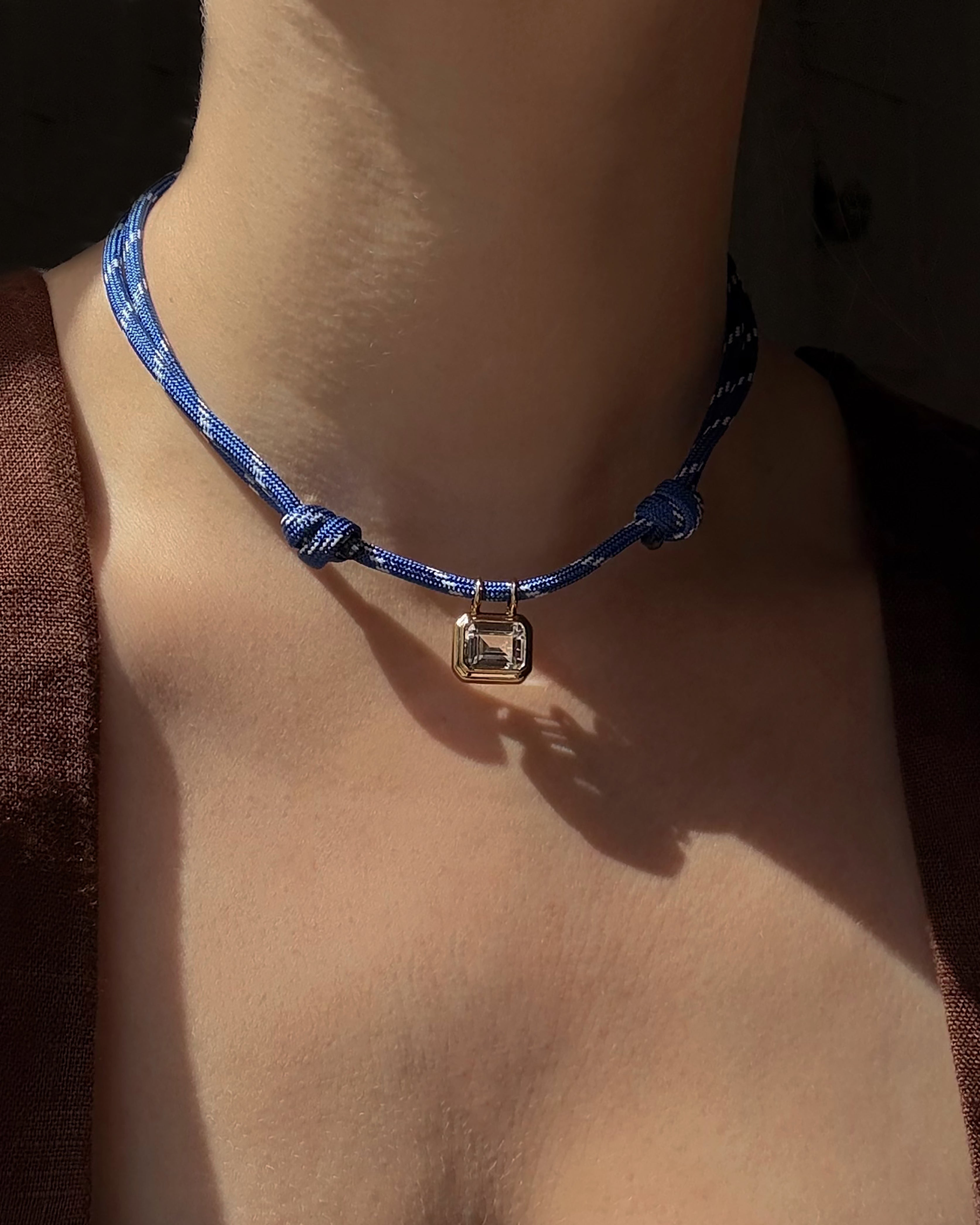 A close-up of a person wearing the *Preorder* Tommy Sapphire Pendant Necklace by Mod + Jo, featuring an adjustable blue cord with knot details and a rectangular silver charm, shown against bare skin and part of a brown garment in warm lighting.