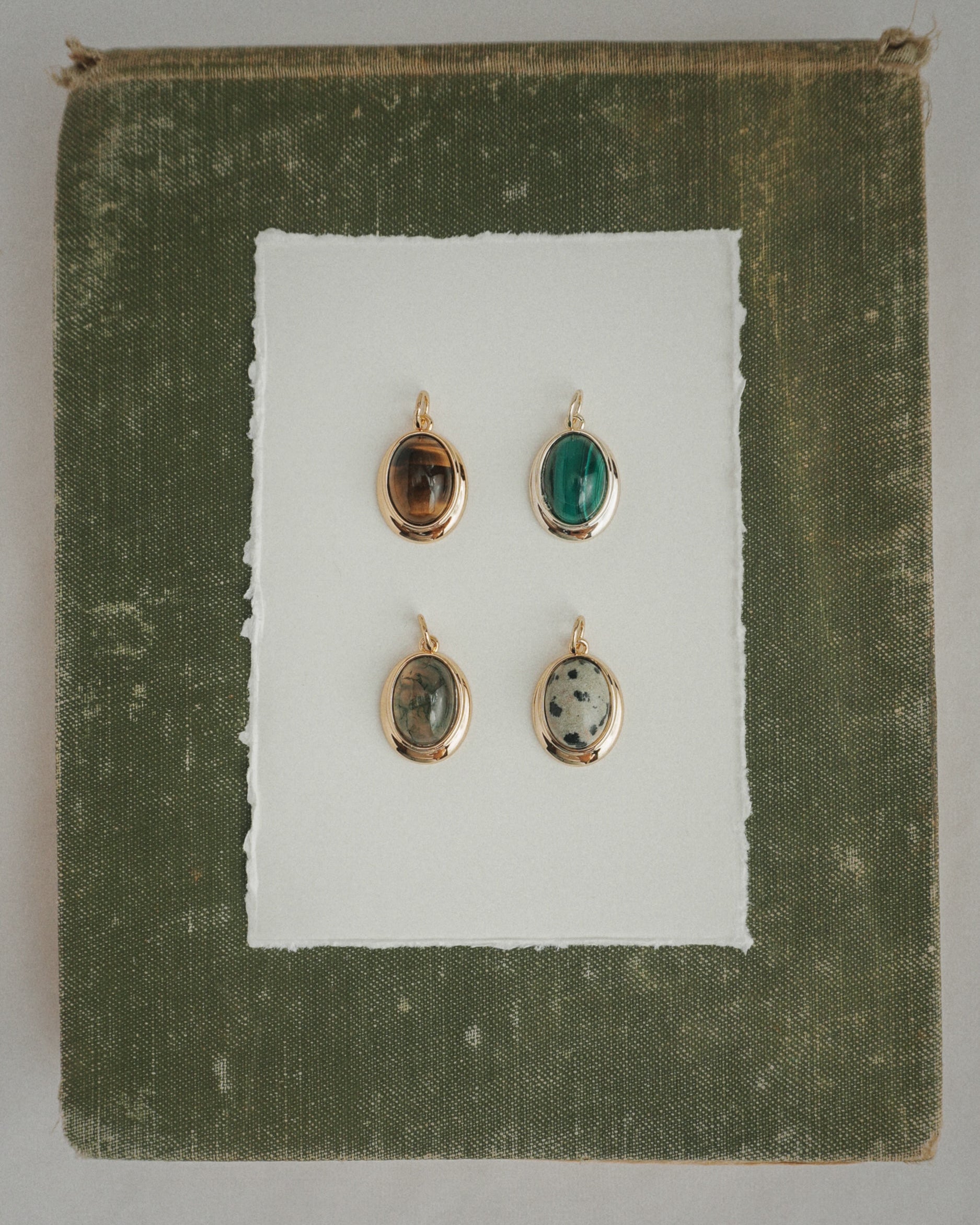 Four Rory Gemstone Charms by Mod + Jo, each featuring a unique oval gemstone with 14k gold over brass edges, are arranged on a sheet of white paper centered against a textured olive green square background. Each charm showcases a different stone—including the striking Dalmatian Jasper—and is thoughtfully crafted for an elegant touch.