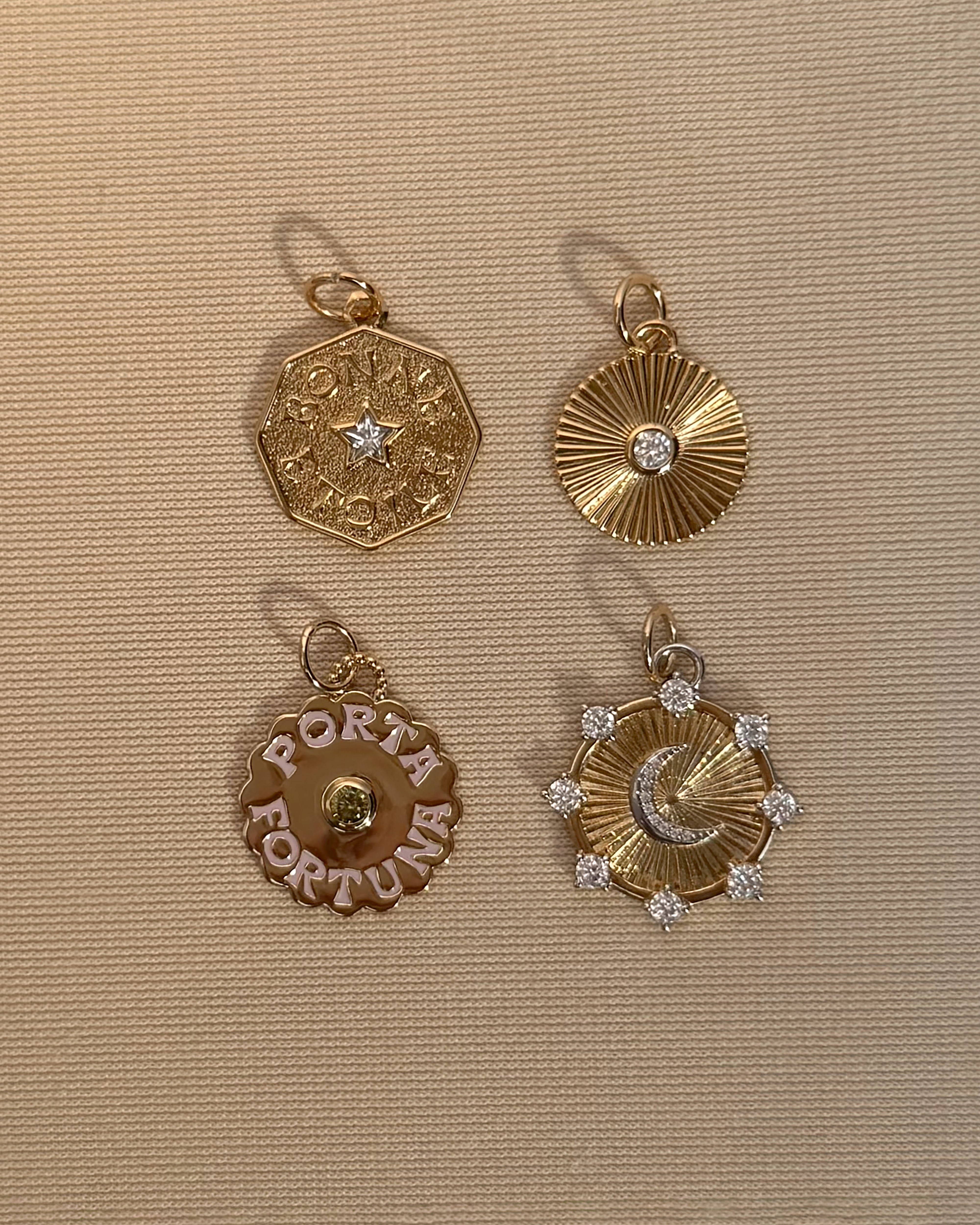 Four gold charms rest on a beige textured background: an octagonal star motif, a ridged circle set with a gem, a "PORTA FORTUNA" gold pendant, and the Lyra Moon Charm by Mod + Jo—a round moon design adorned with gemstones—together creating an enchanting selection of celestial jewelry.