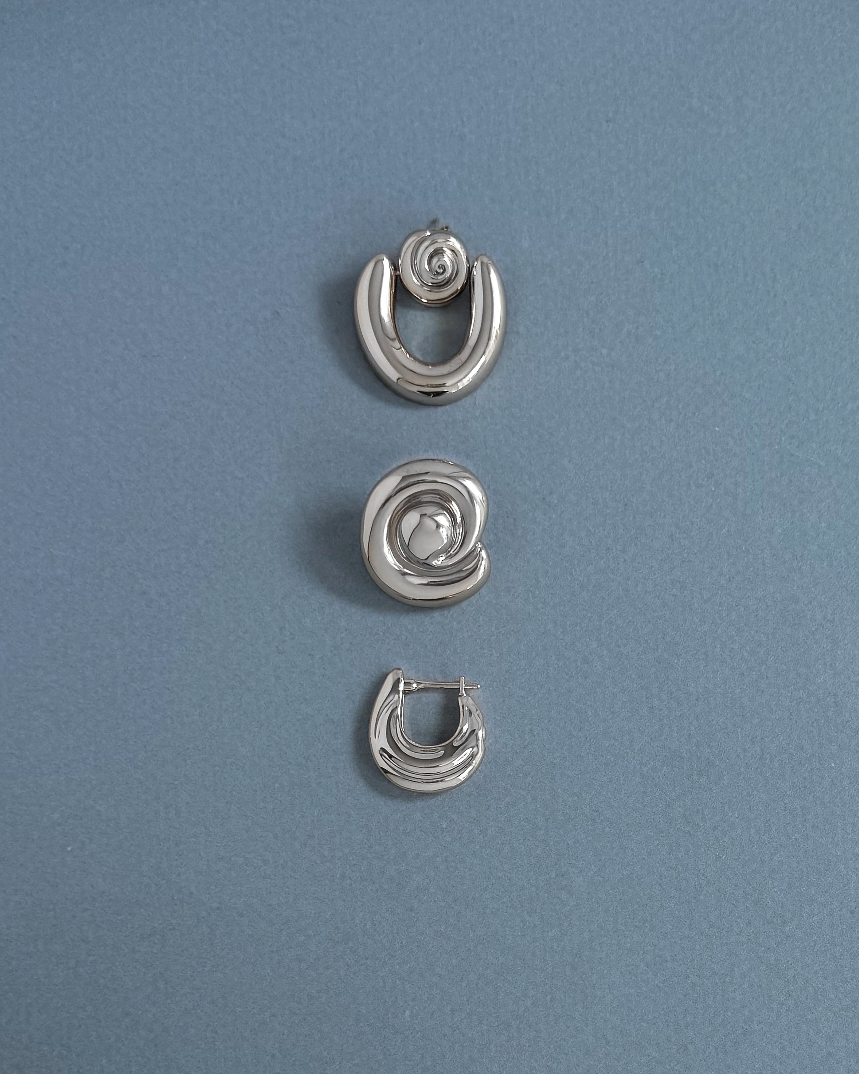 Three silver earrings from Mod + Jo are arranged vertically on a light blue textured background. The top and bottom are crescent-shaped, while the center features the Milley Spiral Statement Studs—an elegant hypoallergenic choice.