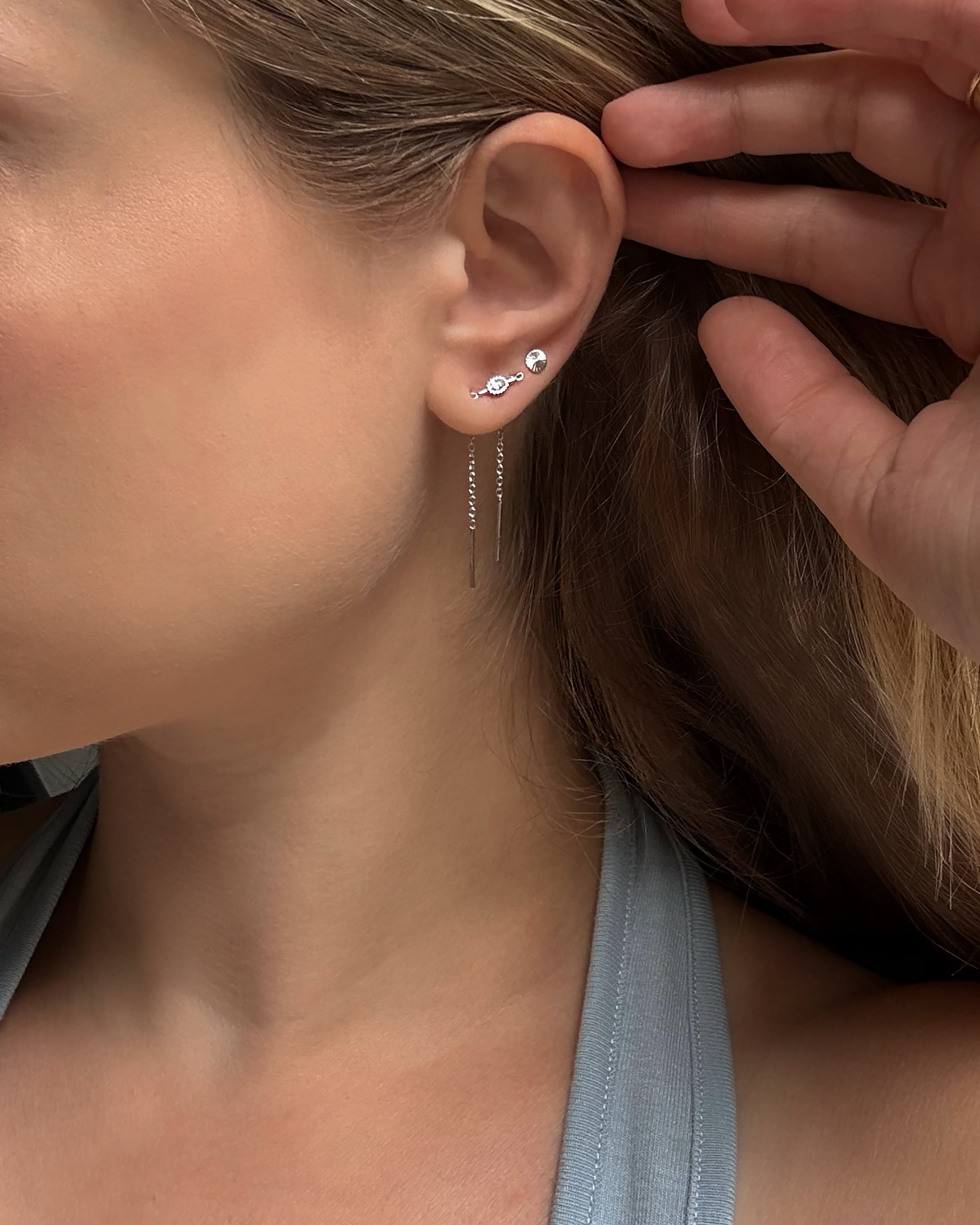 A close-up of a woman’s ear showcases three silver earrings: two cubic zirconia studs on her upper lobe and the Nicole Oval Threader Earring by Mod + Jo—a delicate, rhodium plated chain earring—on her lower lobe. She is dressed in a gray tank top and holds her hair back with one hand.