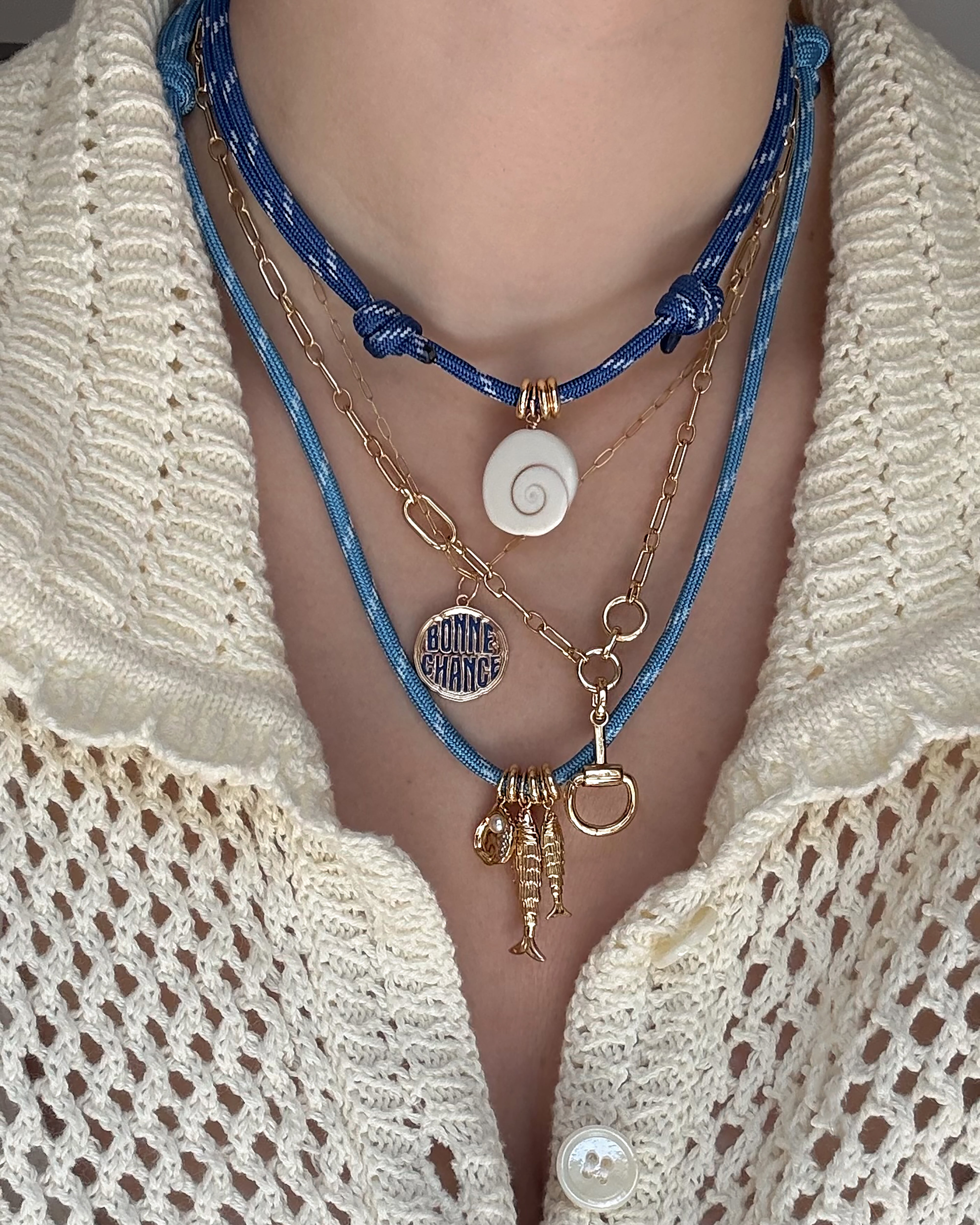 Wearing Mod + Jo's Paige Horsebit Charm Enhancer, layered with blue cords and 14k gold-plated, tarnish-resistant chains adorned with a shell pendant and various charms—including a fish and a round Bonne Chance medallion—paired with a cream crochet button-up top.