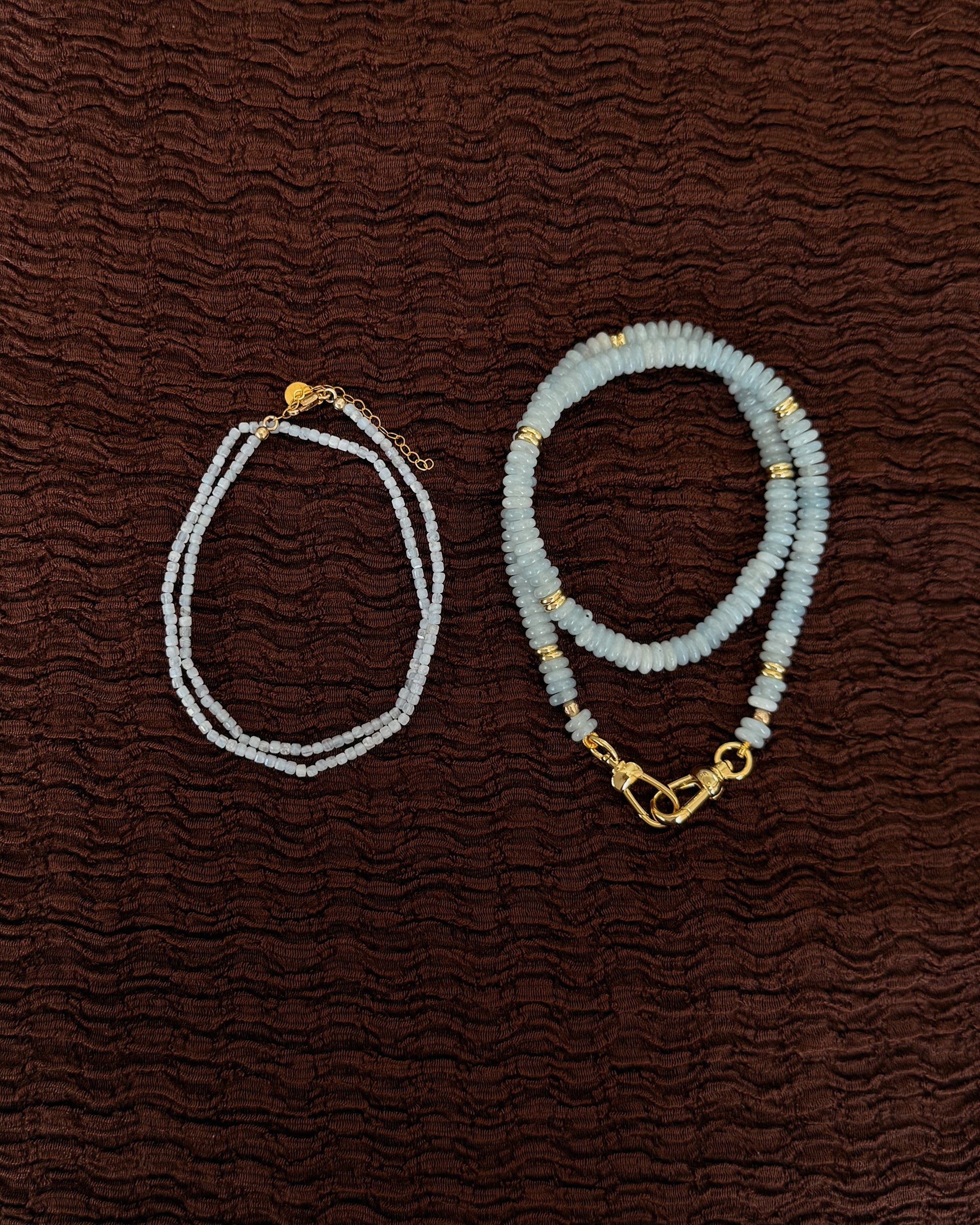 The Mod + Jo Merit Gemstone Necklace is displayed on textured brown fabric, featuring vibrant blue gemstone beads with gold accents and a secure clasp.