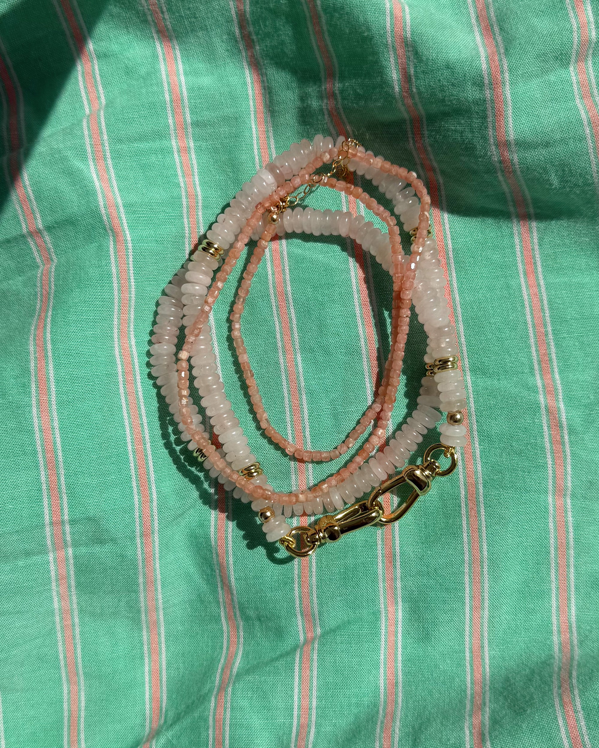 The Sonia Gemstone Necklace by Mod + Jo features pink and white genuine gemstone beads with gold clasps, coiled on green striped fabric and shown in natural sunlight—a beautiful piece of handmade jewelry.