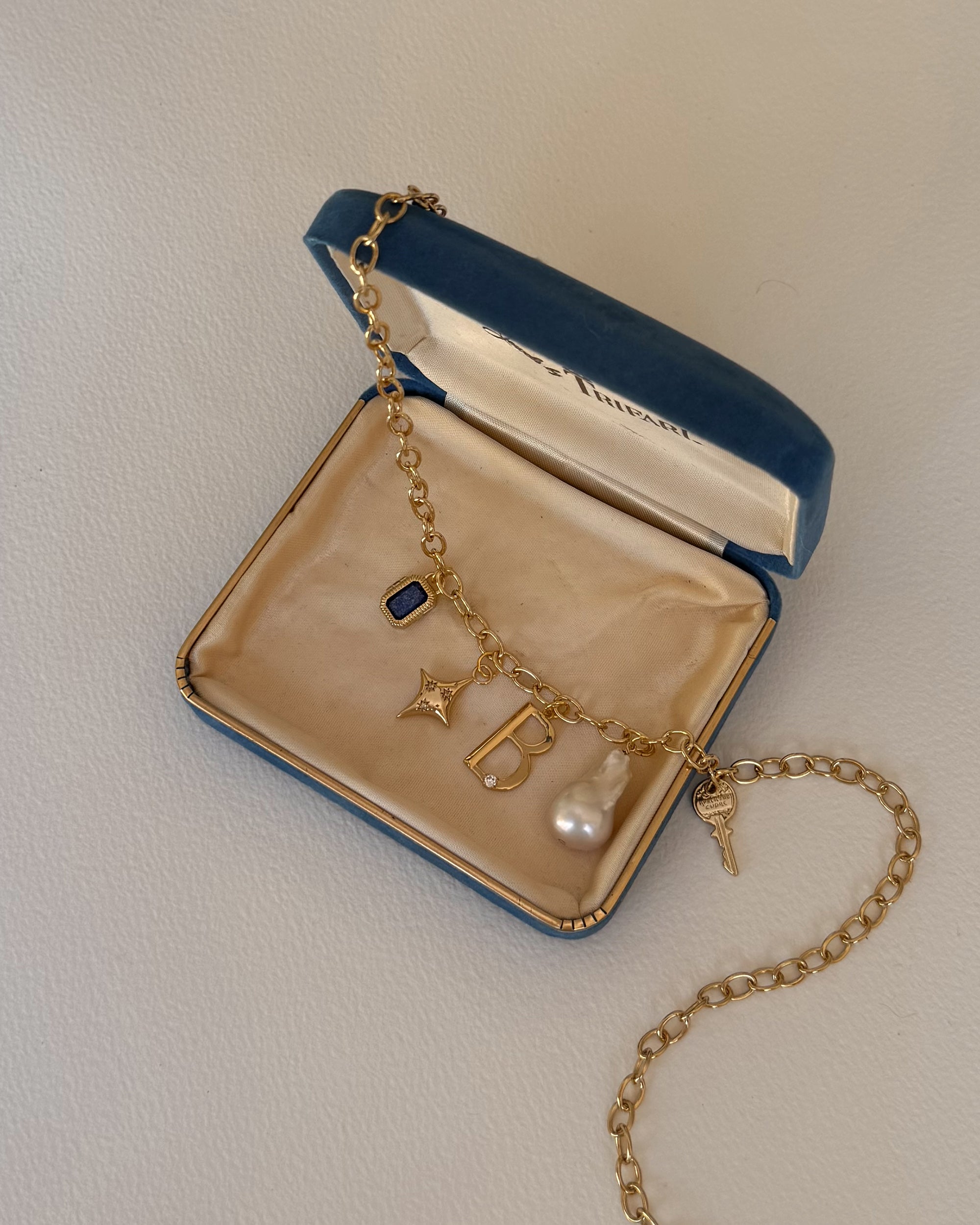 The Eloise Letter Charm bracelet by Mod + Jo, featuring gold plating, a blue gemstone, star, initial charm, pearl, and key, is displayed in an open blue velvet jewelry box with beige lining on a light surface.