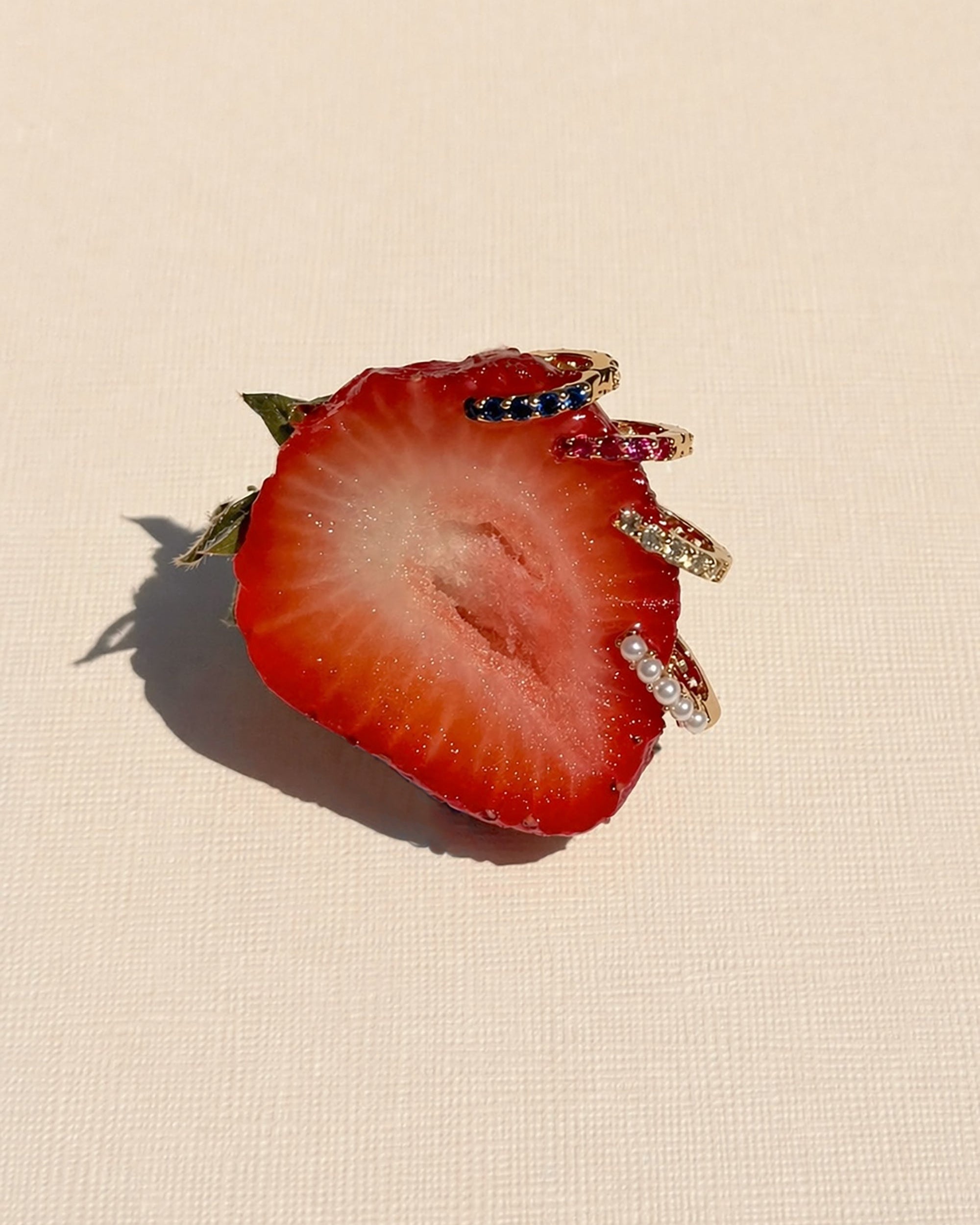 A sliced strawberry half displays the Mod + Jo Harlow Reversible Birthstone Huggies, adorned with colorful gemstones and pearls, set against a textured light background.