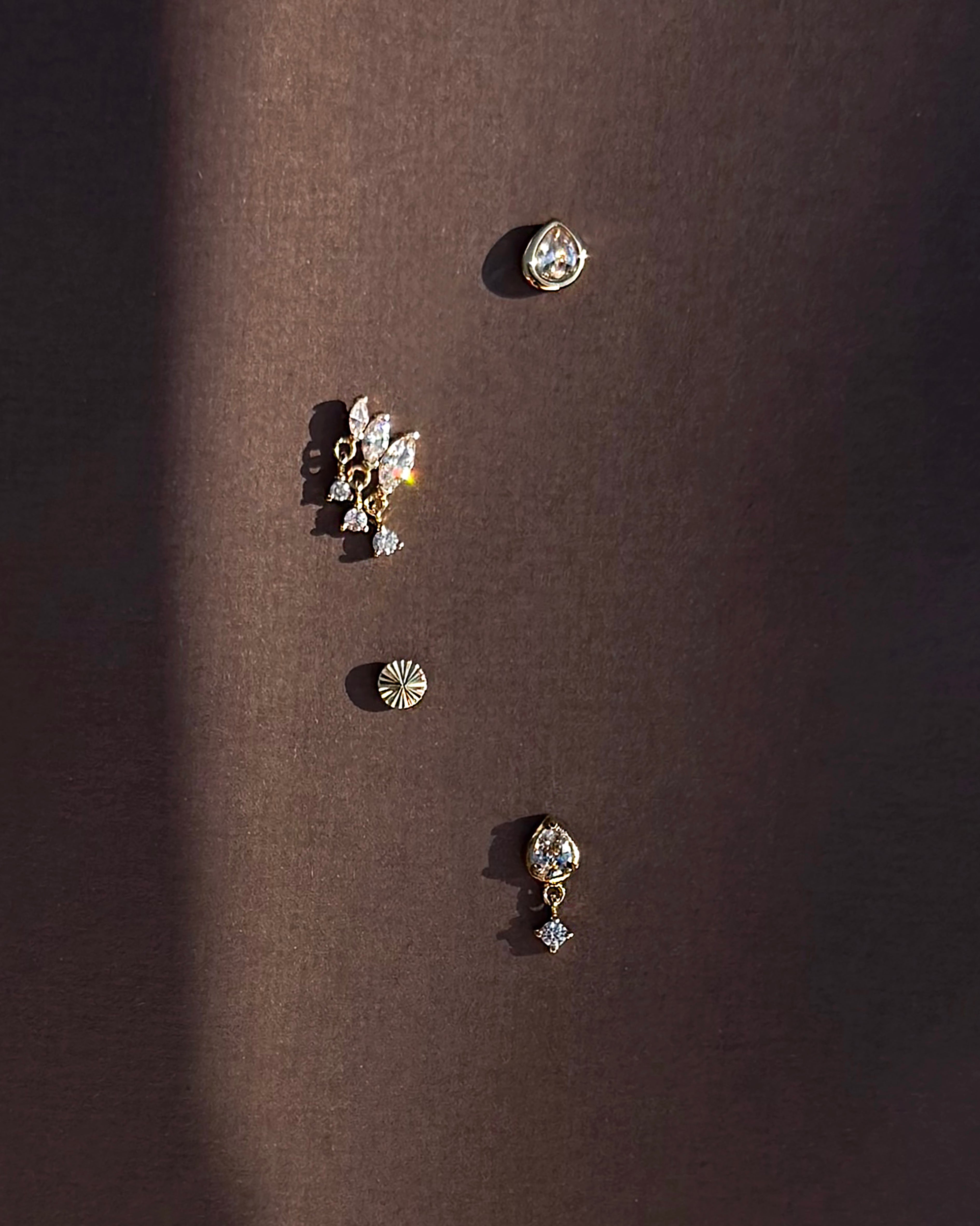 Four unique earrings from Mod + Jo, including the Joni Bezel Studs, are arranged vertically on a dark brown background. They showcase gold- and silver-toned designs in various shapes with sparkling stones like cubic zirconia, all casting soft shadows.