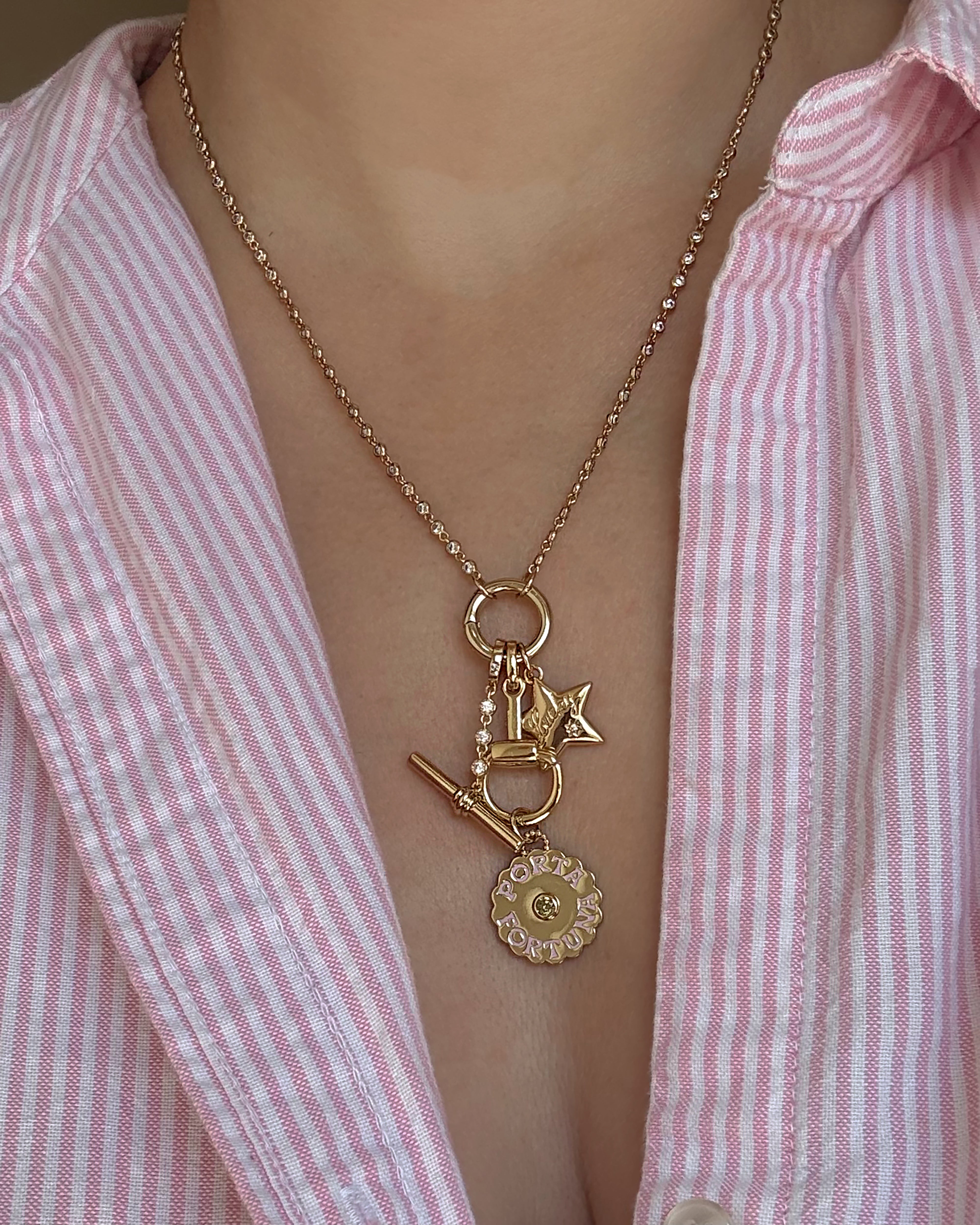 Worn over a pink and white striped shirt, the Mod + Jo Livia Tennis Toggle Charm necklace features gold plating and showcases a key, star, and circular CZ charm engraved with "LOVE," all adorned with sparkling rhinestones.