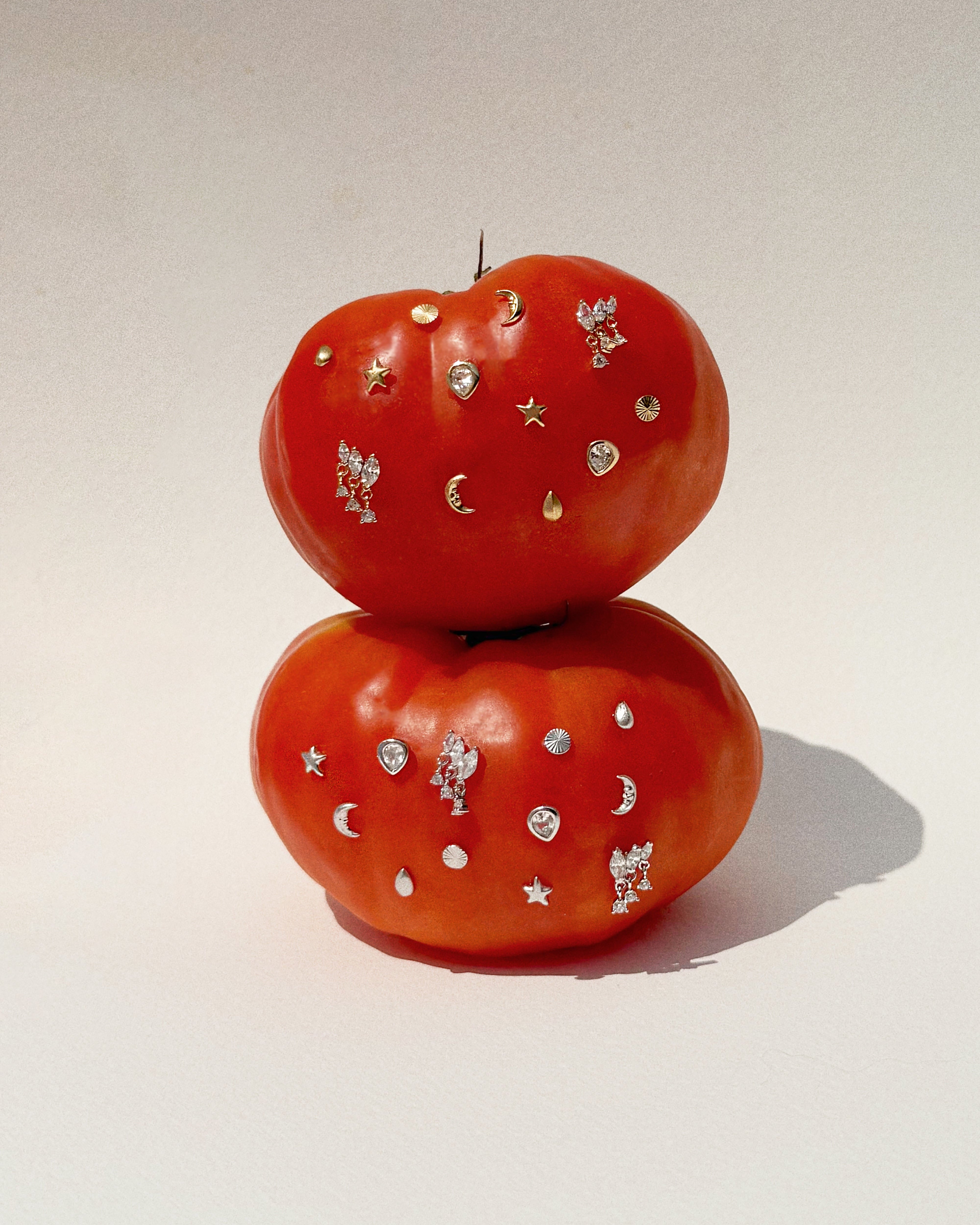 Two red tomatoes stacked on a pale surface, adorned with small metallic stickers in star, moon, drop, and other shapes—evoking the delicate shine of the Gabby Marquise Dangle Studs by Mod + Jo.
