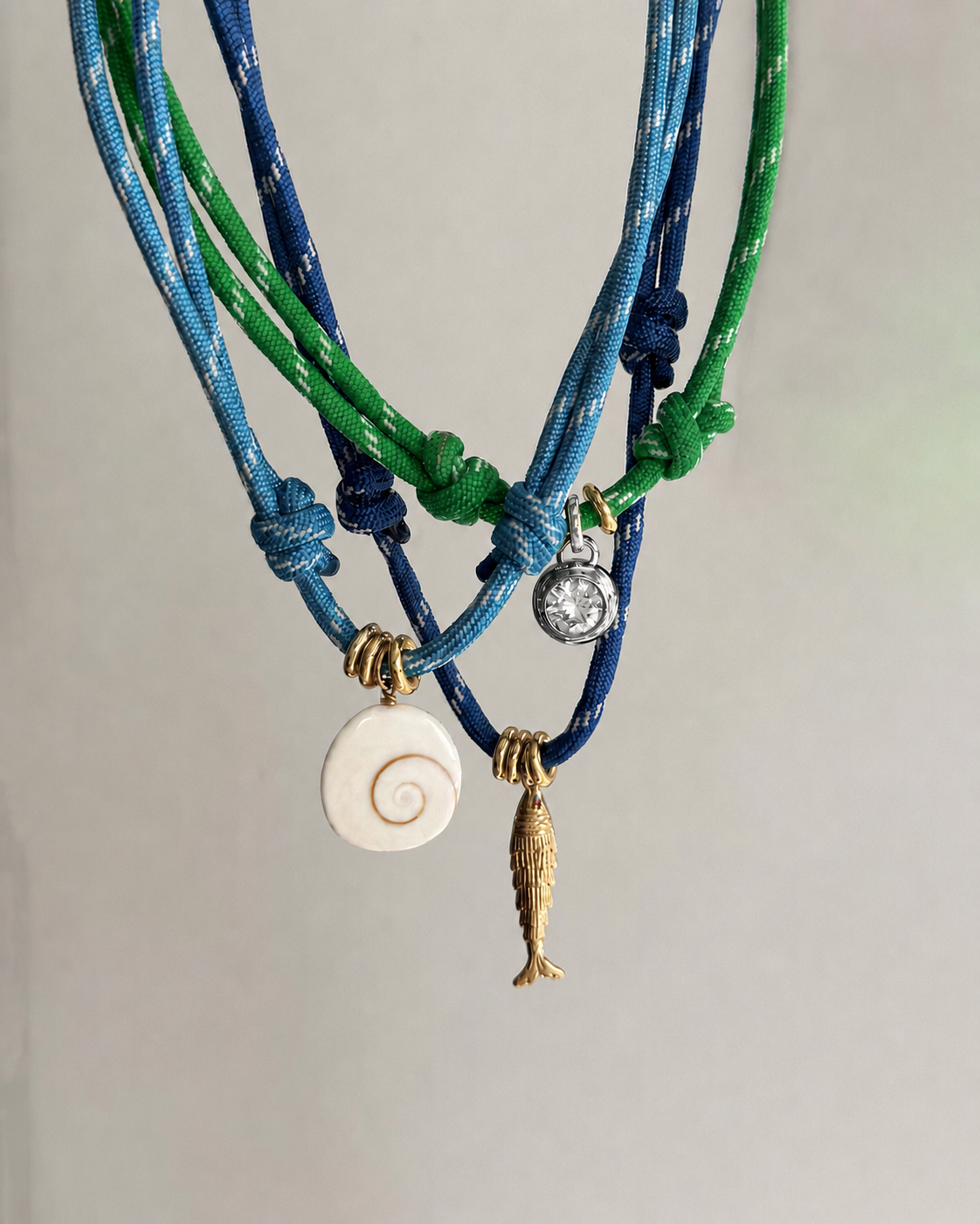 Three cord necklaces—two blue, one green—hang side by side. Charms include a white spiral shell, silver coin, gold fish, and Mod + Jo’s Tove Sapphire Bezel Charm with a lab-grown white sapphire. The background is light-colored.