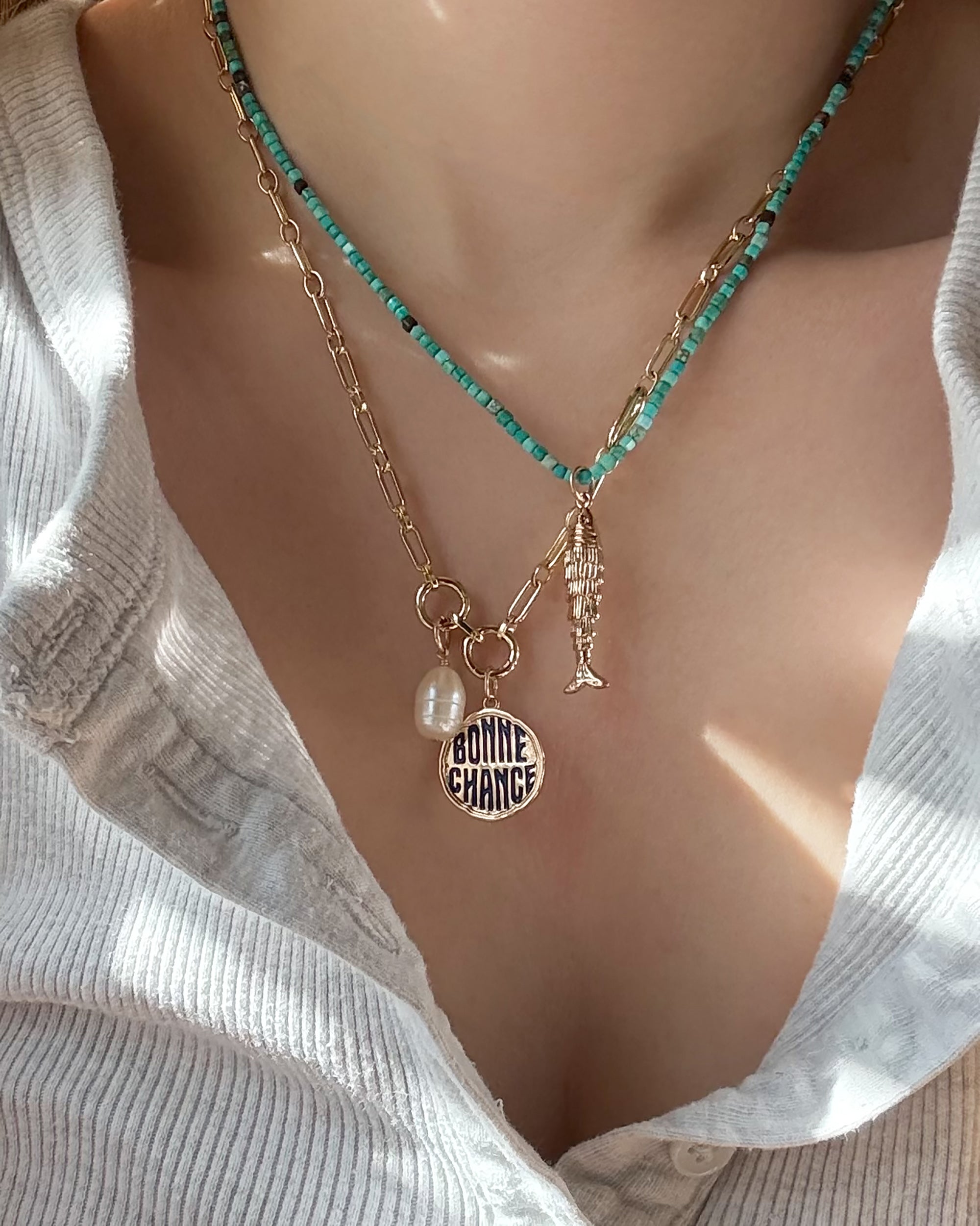 A close-up of a person wearing a white ribbed top and hand-crafted necklaces: the Mod + Jo Merit Gemstone Necklace, a gold chain with a pearl, a fish charm, and a round pendant reading BONNE CHANCE.
