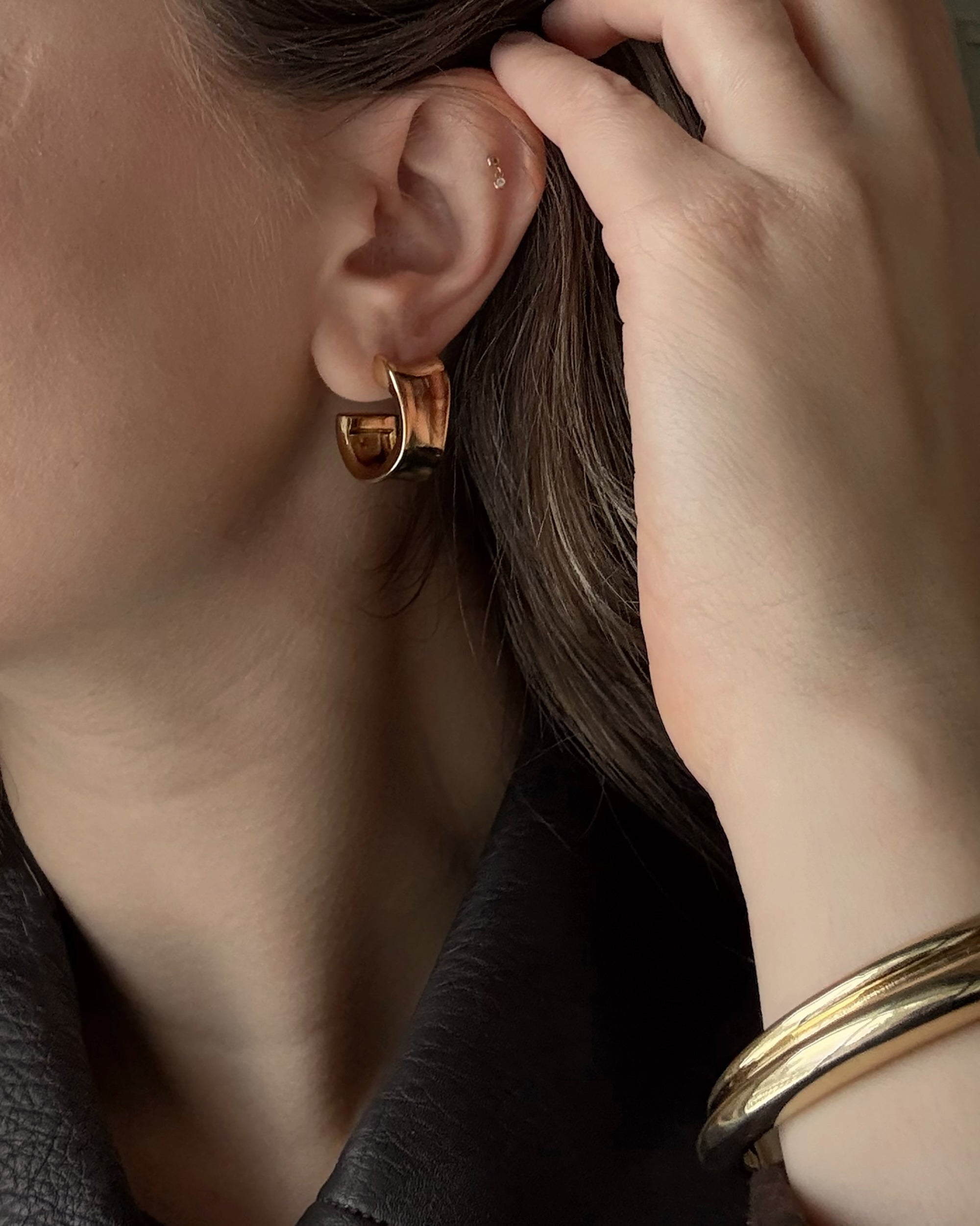 A close-up of a person with light skin wearing a gold hoop earring and the Leila Dome Cuff Bracelet by Mod + Jo, with their hand touching their hair while dressed in a dark, textured top.