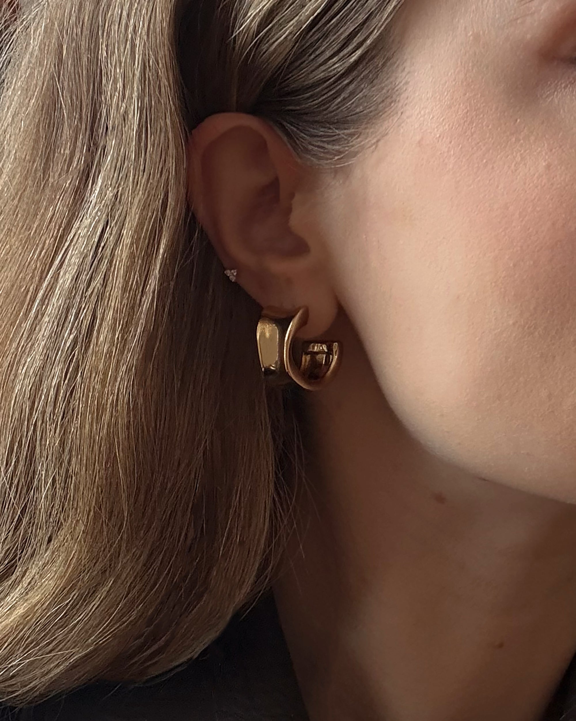 Close-up of a person's ear wearing Zara Volume Hoops and a small stud earring. The person has light brown hair, fair skin, and the background is softly blurred.